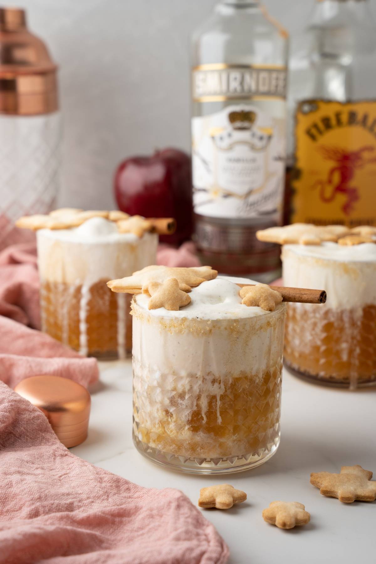Three creamy cocktails in textured glasses, each topped with foam, cookies, and a cinnamon stick—perfect for enjoying an Apple Pie Cocktail or trying a new autumn cocktail recipe. Liquor bottles sit behind for extra inspiration.