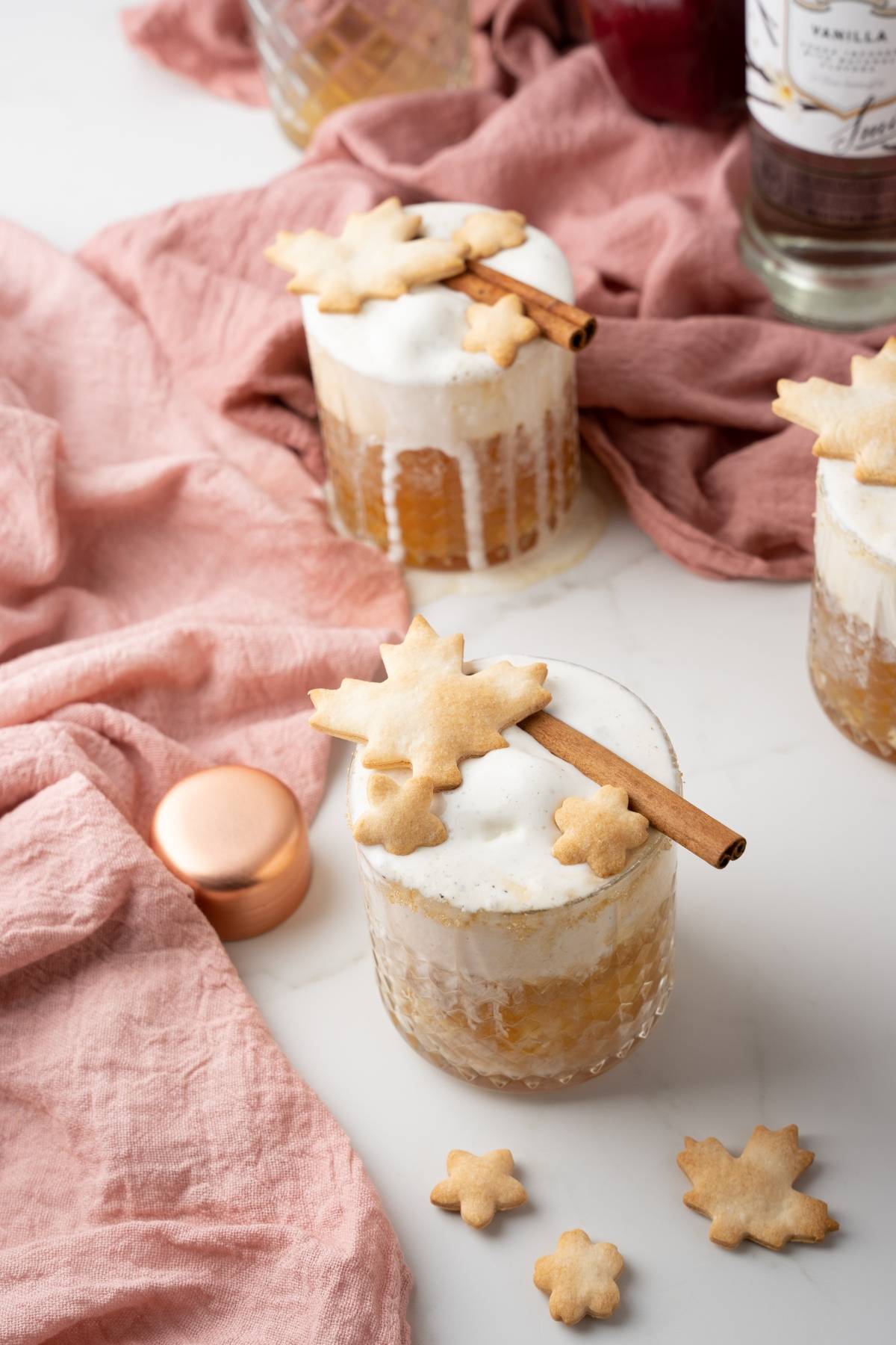 Two cocktails with foamy tops, garnished with cinnamon sticks and leaf-shaped cookies on a pink cloth—an inviting Apple Pie Cocktail perfect for any autumn cocktail recipe collection.
