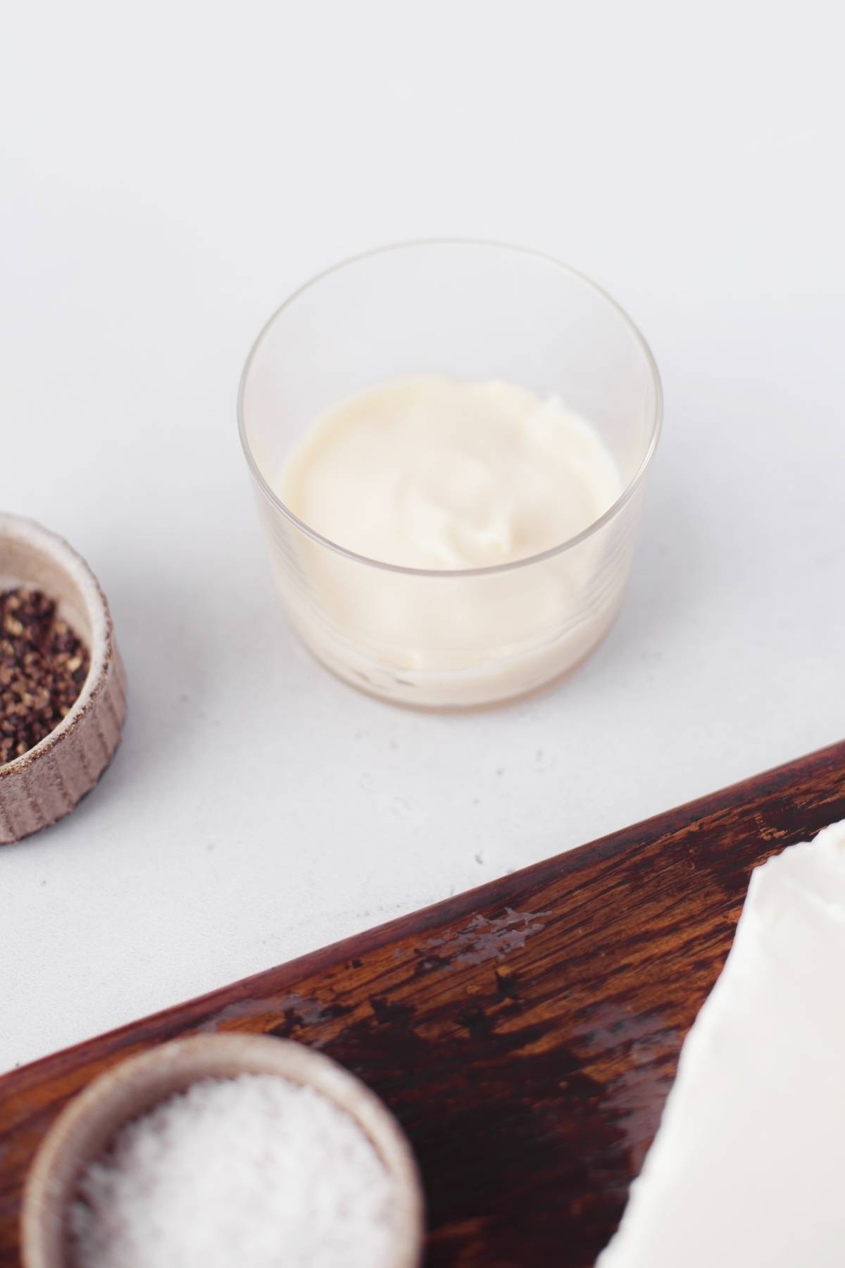 A glass cup of mayonnaise next to small bowls of salt and pepper on a white surface, ready for an easy appetizer dip recipe or a classic hot artichoke appetizer.