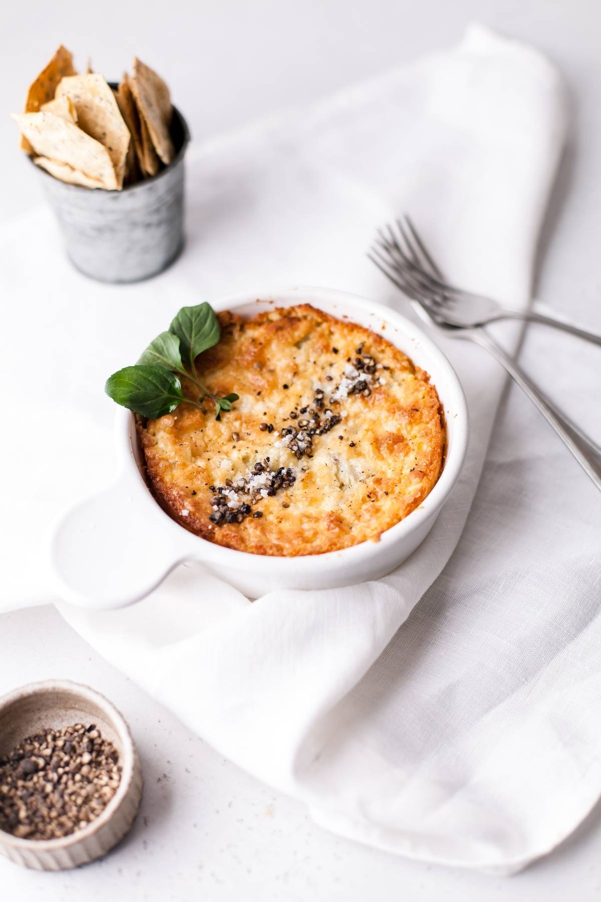 A baked artichoke dip in a white dish, garnished with herbs, sits beside crackers, forks, and a small bowl of pepper—an easy appetizer dip recipe made with cream cheese.