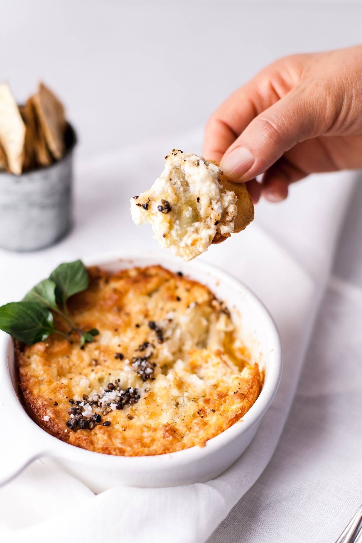 A hand dips a cracker into a cheesy baked artichoke dip in a white dish, garnished with herbs—an irresistible appetizer made with cream cheese.