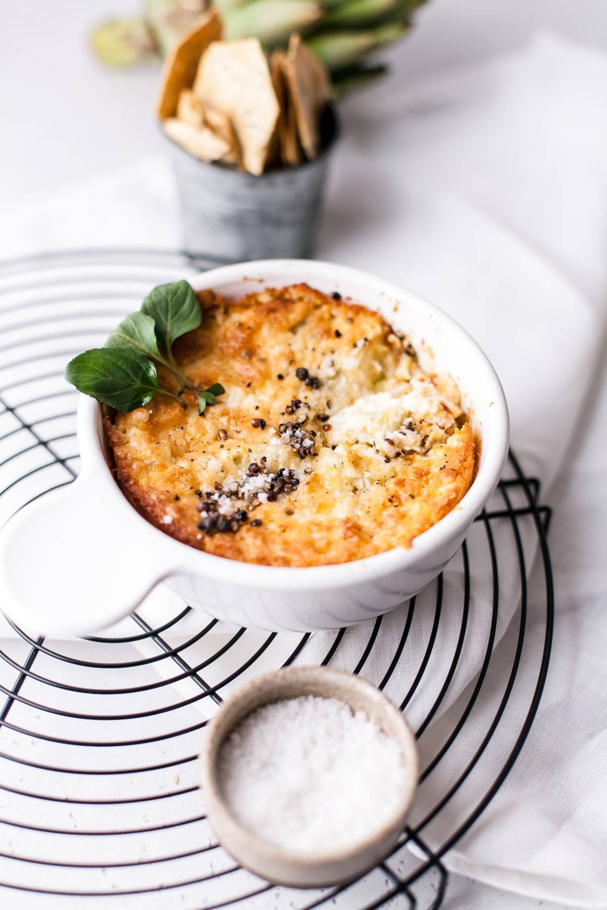 Baked artichoke dip in a white ramekin, garnished with herbs, beside a bowl of salt and crackers in the background—an easy appetizer dip recipe perfect for any gathering.