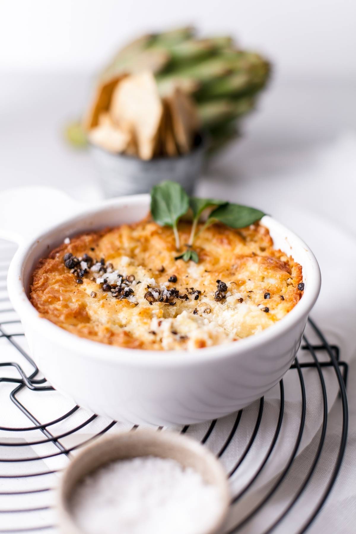 Golden baked appetizer made with cream cheese in a white bowl, garnished with herbs; artichokes and salt nearby.