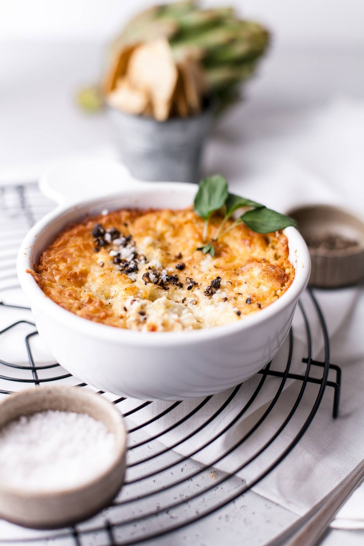 Baked artichoke dip topped with herbs and cheese in a white bowl on a cooling rack, with salt and chips nearby—an easy appetizer dip recipe perfect for any gathering.