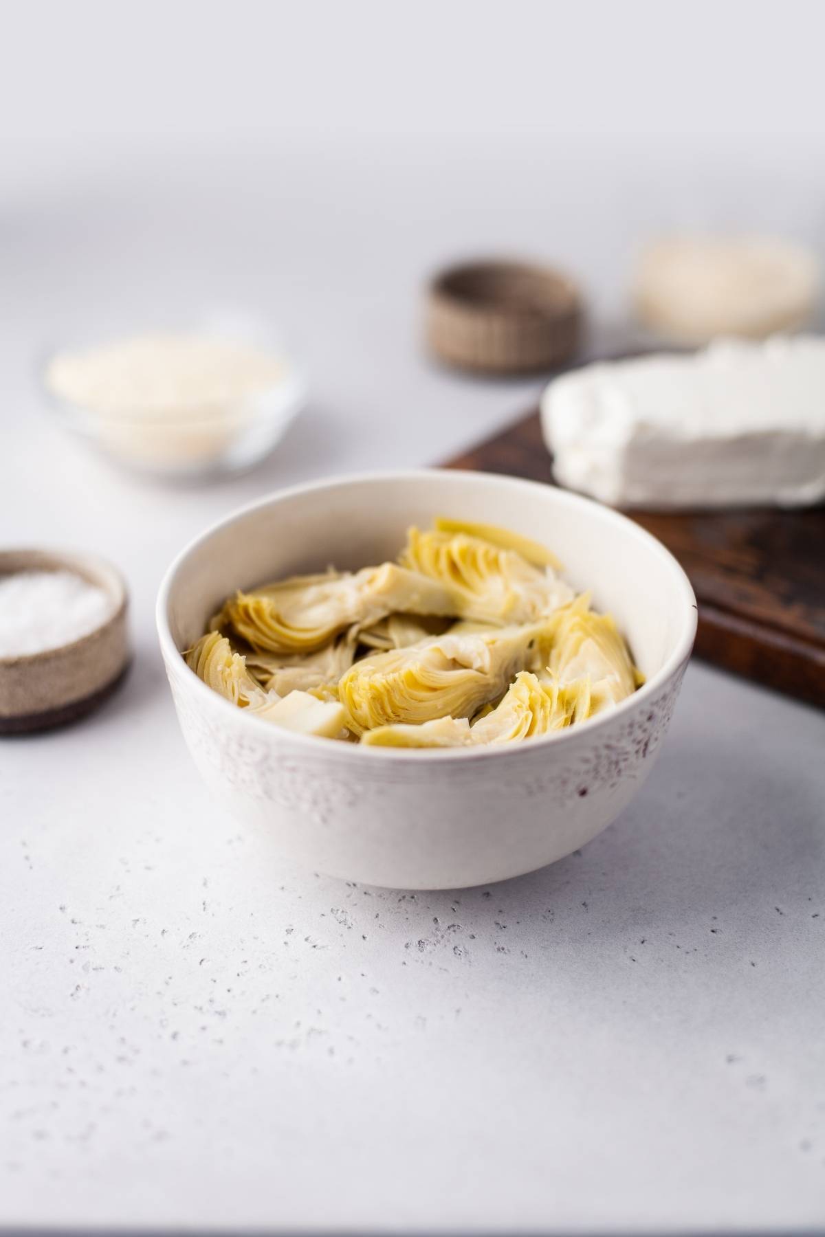A bowl of artichoke hearts sits on a light surface with cheese and salt in the background, perfect for using in your favorite recipe with artichoke hearts or as a base for a creamy artichoke dip.