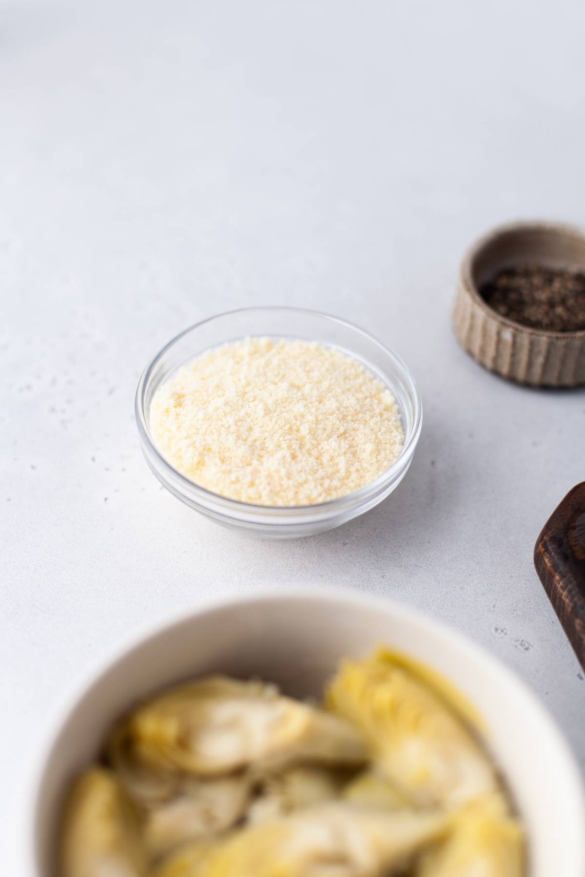 A small glass bowl filled with grated Parmesan cheese on a light surface, ready to be used in an artichoke dip or another delicious appetizer made with cream cheese, with other ingredients nearby.