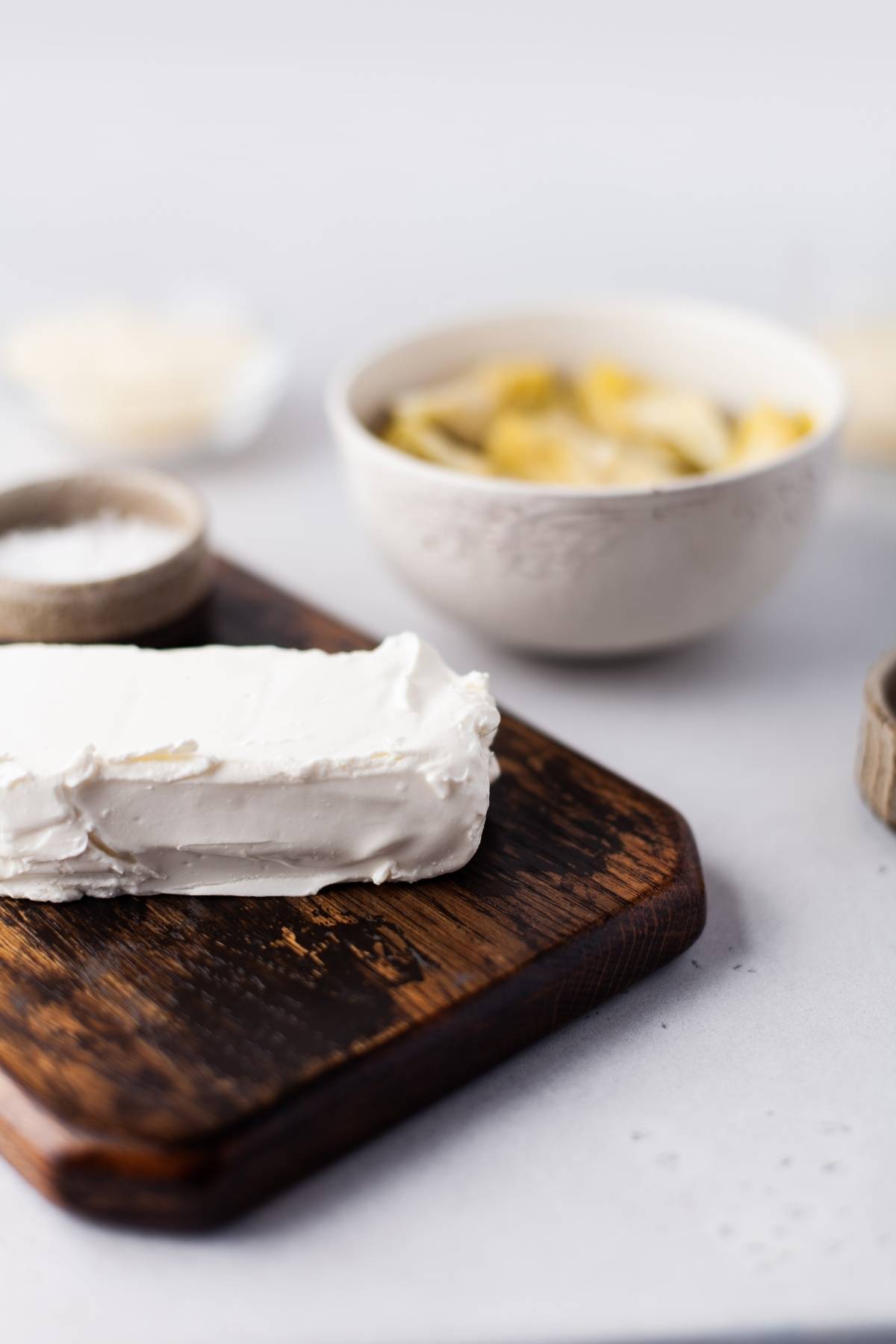 A block of cream cheese on a wooden board with a bowl of artichoke hearts in the background—perfect for creating an easy appetizer dip recipe.