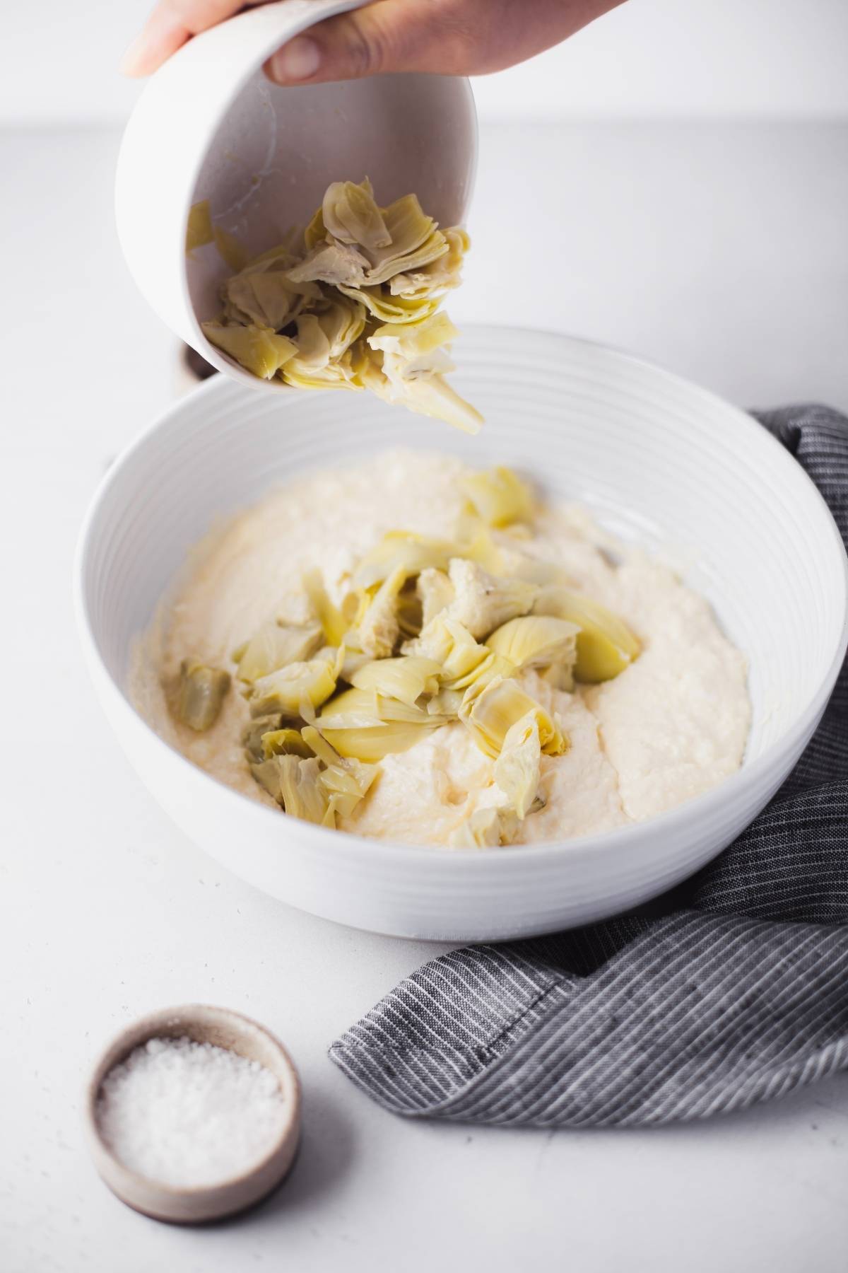 A hand pours chopped artichokes into a white bowl of creamy dip, creating an easy appetizer dip recipe. A striped napkin and salt sit nearby, ready to complement this irresistible recipe with artichoke hearts.
