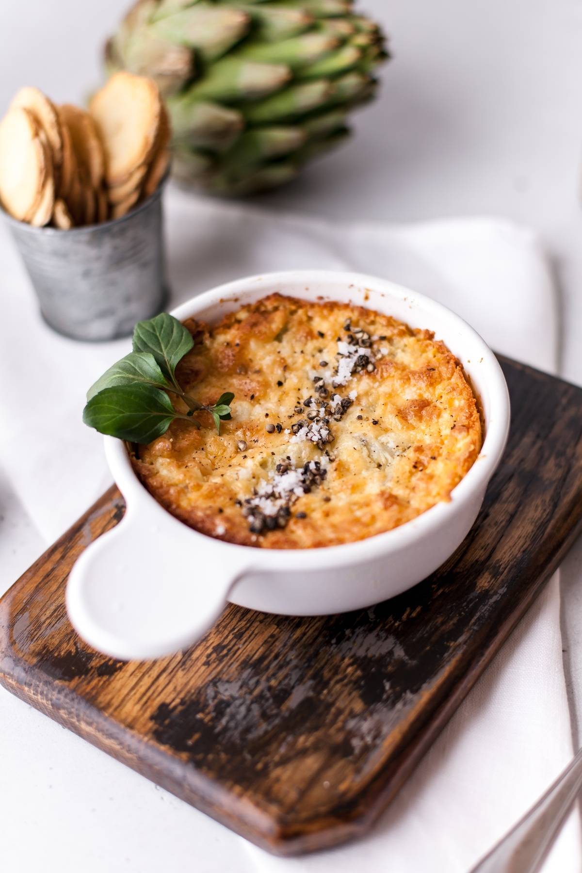 A baked cheesy artichoke dip in a white ramekin, garnished with herbs, on a wooden board with crackers nearby—an irresistible appetizer made with cream cheese.