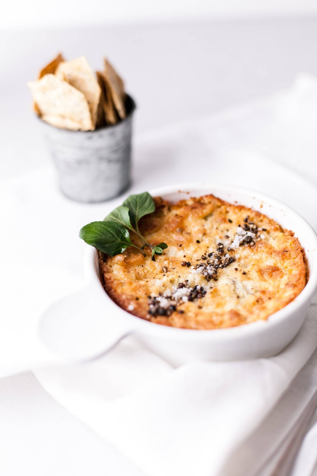 Baked artichoke dip in a white dish, garnished with herbs, with a cup of crackers in the background—an easy appetizer dip recipe perfect for any gathering.