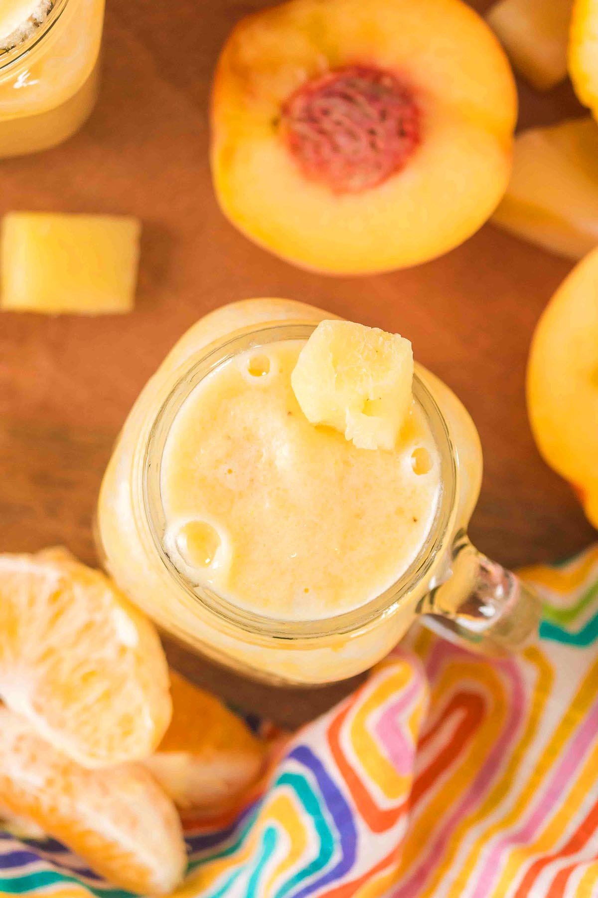 A creamy Pineapple Peach Smoothie in a glass jar, topped with pineapple, surrounded by peach and orange slices, and set on a colorful cloth.