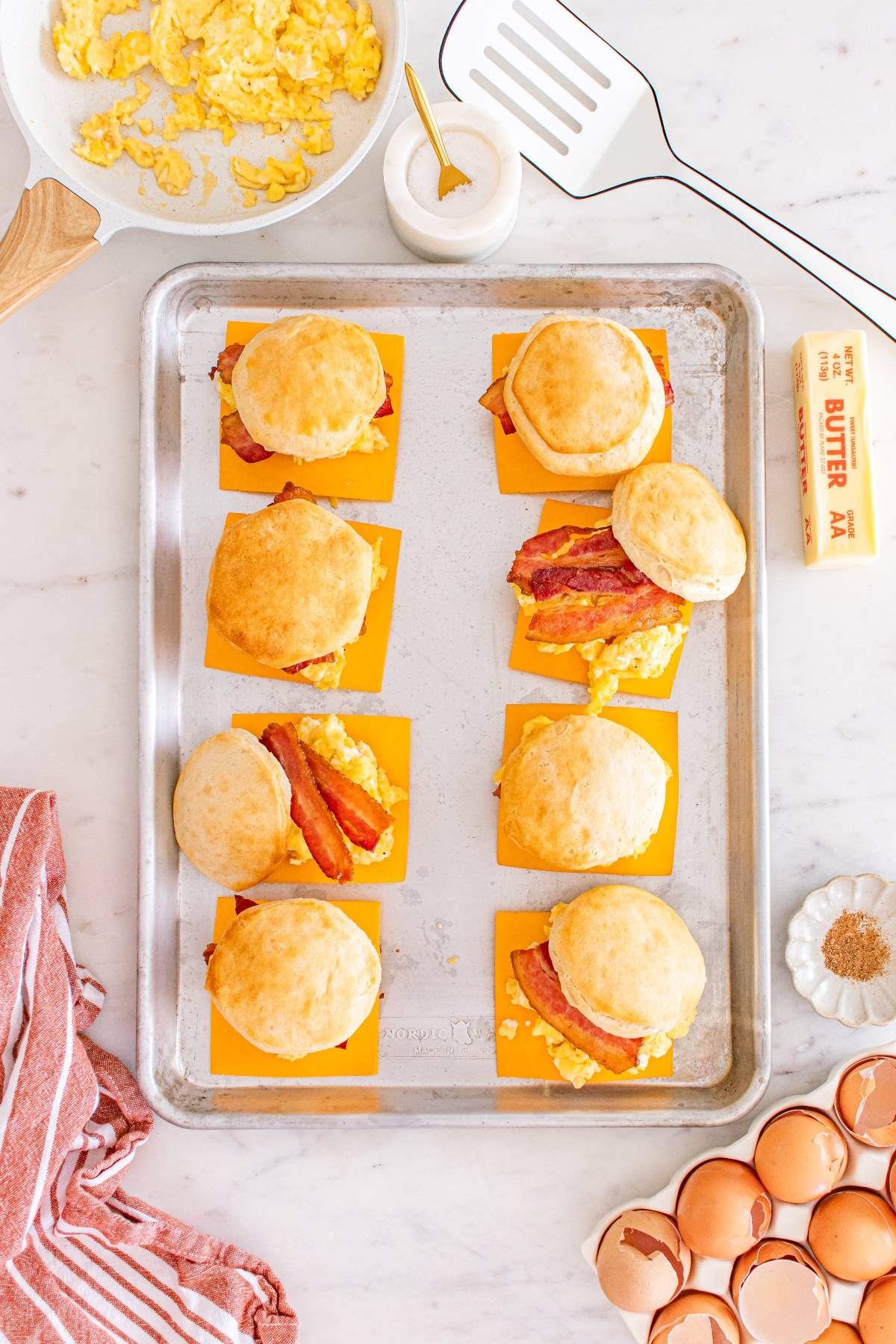 A baking tray with six biscuit breakfast sandwiches with egg, cheese, and bacon on a marble countertop.