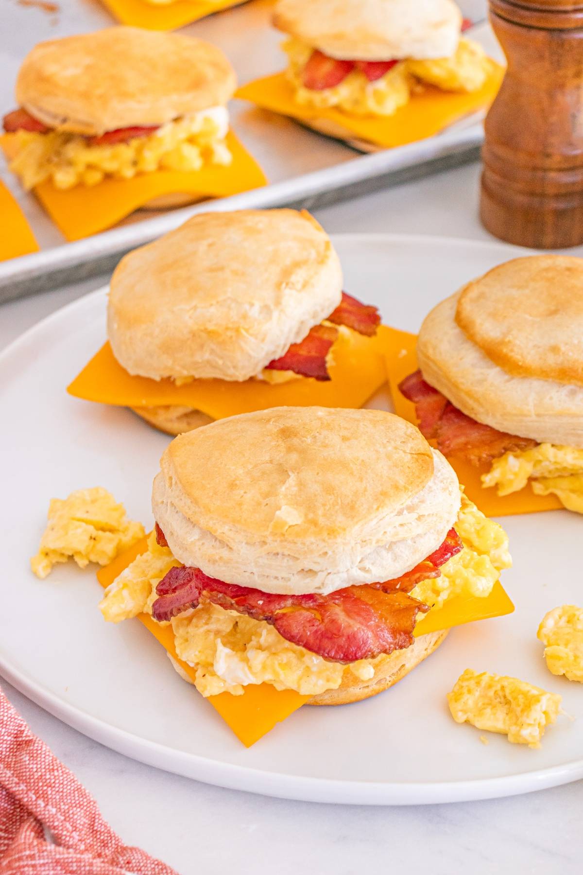 Biscuits filled with scrambled eggs, cheddar cheese, and bacon on a white plate, with extras in the background.