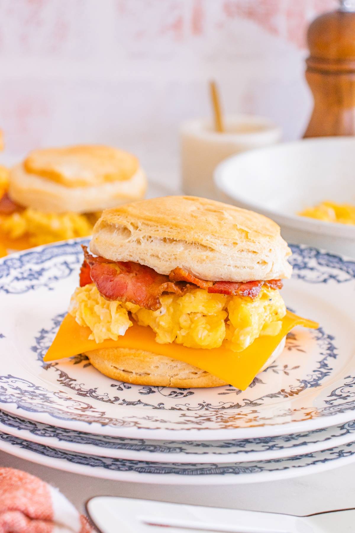 Breakfast sandwich with bacon, scrambled eggs, and cheddar cheese on a biscuit, served on a decorative plate.