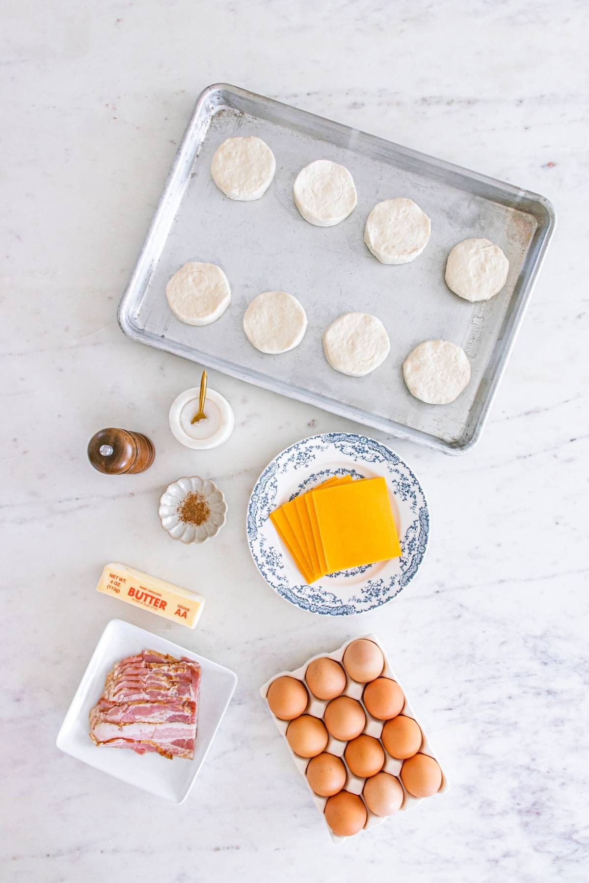 Baking sheet with biscuit dough, cheese slices, eggs, bacon, butter, and seasonings on a marble surface.