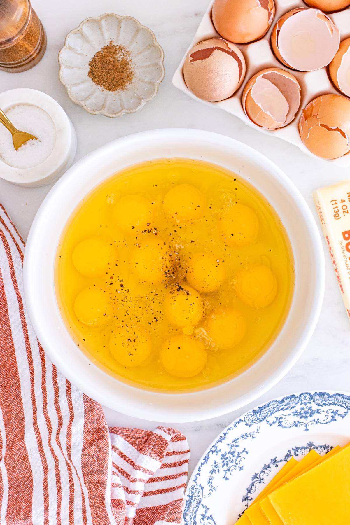 A bowl of cracked eggs with pepper, surrounded by egg shells, cheese slices, and seasonings on a counter.