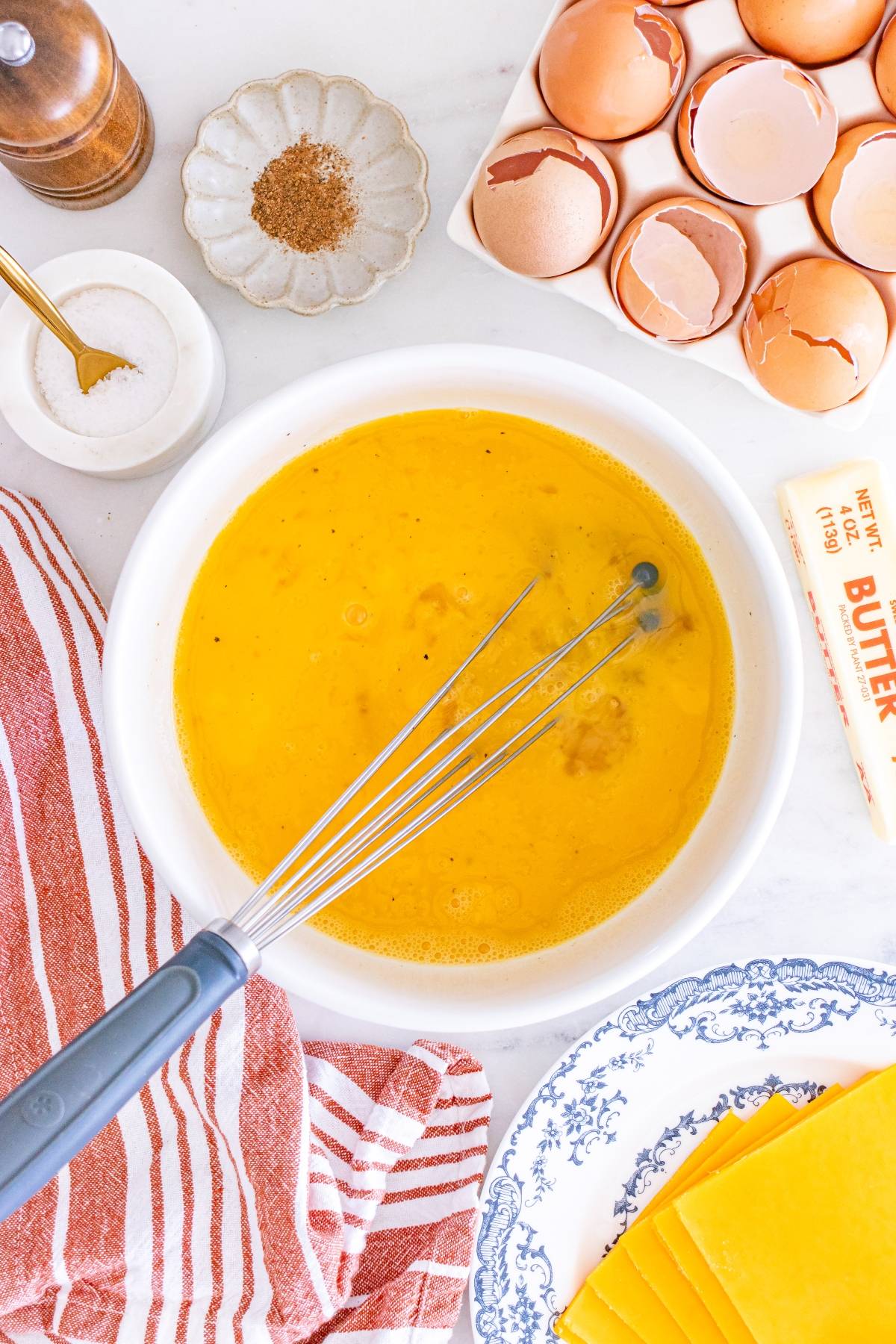 Whisking eggs in a bowl with butter, cheese slices, spices, and cracked eggshells nearby on a countertop.