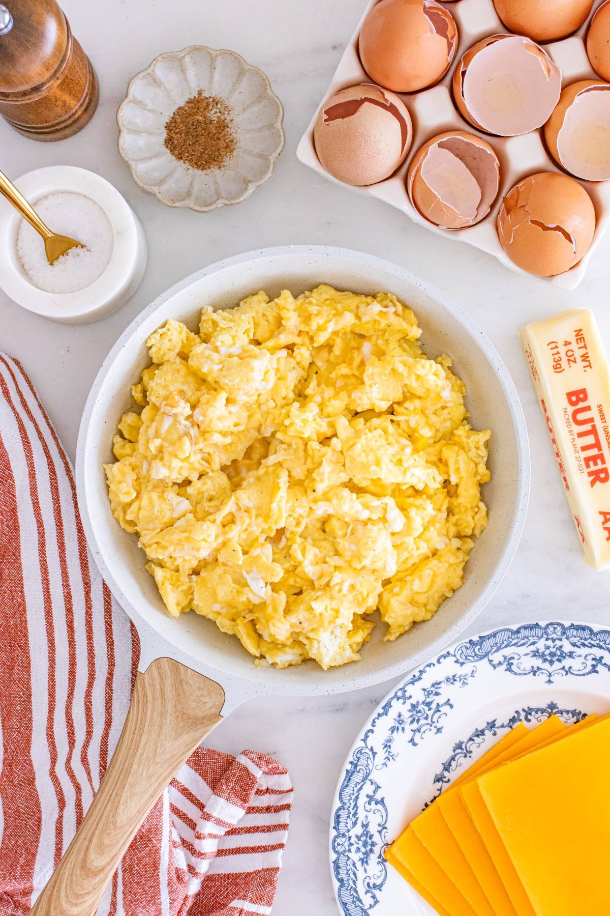 A pan of scrambled eggs surrounded by butter, cheese slices, eggshells, salt, and seasoning on a counter.