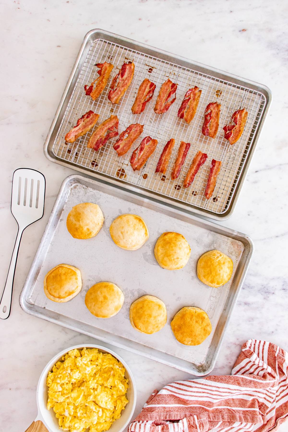 Baked bacon, biscuits on trays, scrambled eggs in a bowl, and a spatula on a marble countertop.
