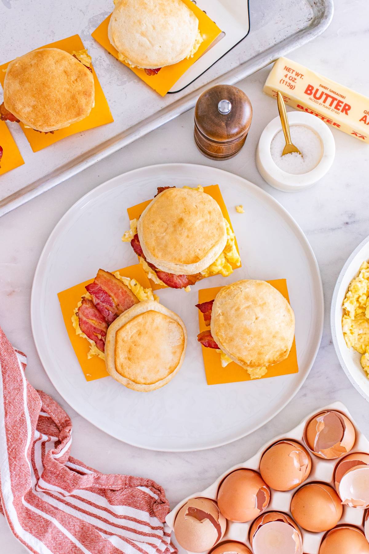 Three breakfast sandwiches with egg, bacon, and cheese on biscuits, surrounded by eggs, butter, and salt.