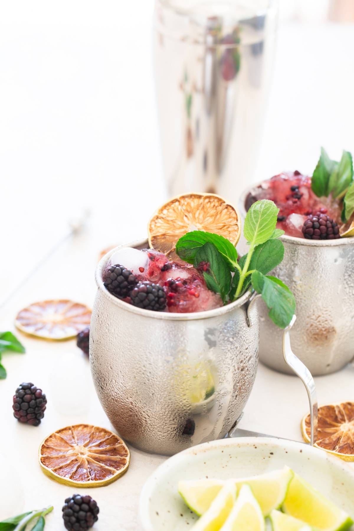Two metal cups with a refreshing blackberry cocktail recipe, garnished with mint, blackberries, lime slices, and dried citrus.