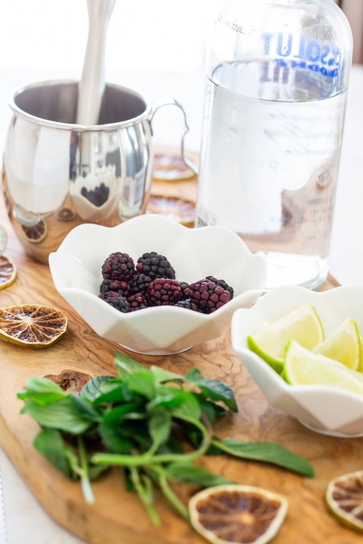 Blackberries, lime wedges, mint, dried citrus, a metal cup, and a bottle of vodka on a wooden board—perfect for a blackberry cocktail recipe or if you're wondering how to make a Moscow mule with a fruity twist.