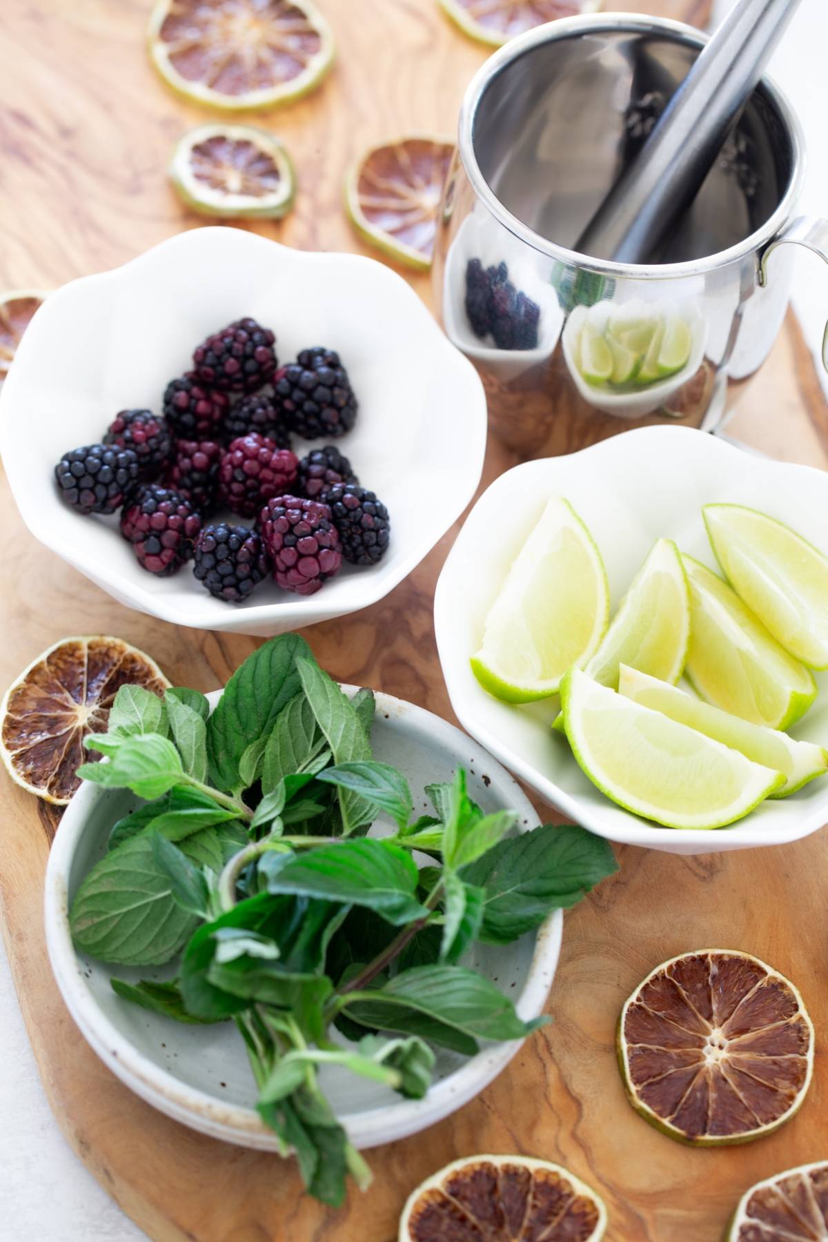 Bowls of blackberries, lime wedges, and mint on a wooden board with dried citrus slices and a metal muddler—perfect for crafting a refreshing blackberry cocktail recipe with vodka.