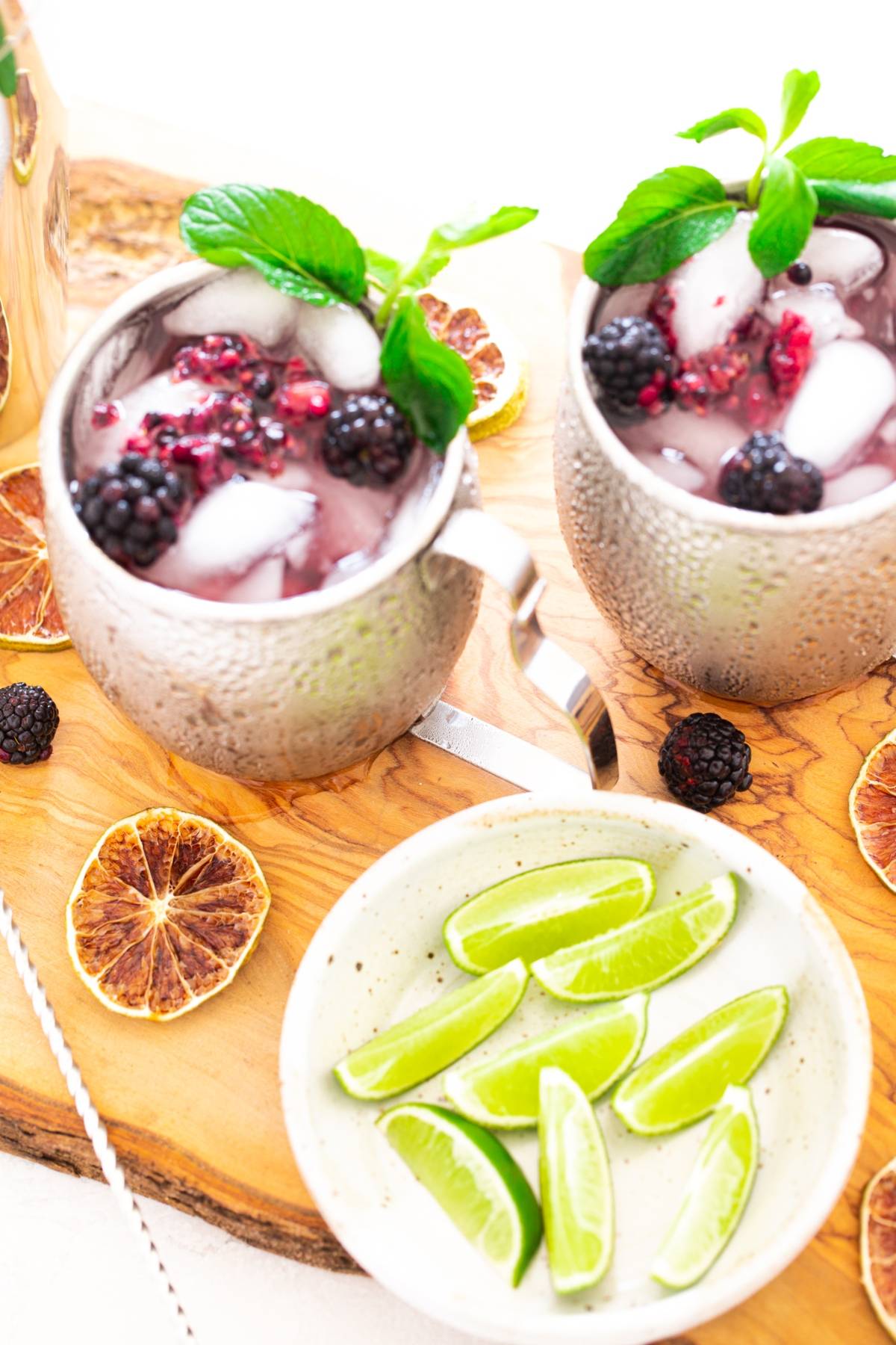 Two metallic mugs with iced berry drinks and mint, perfect for a Blackberry Moscow mule, sit beside a plate of lime wedges and dried citrus slices.