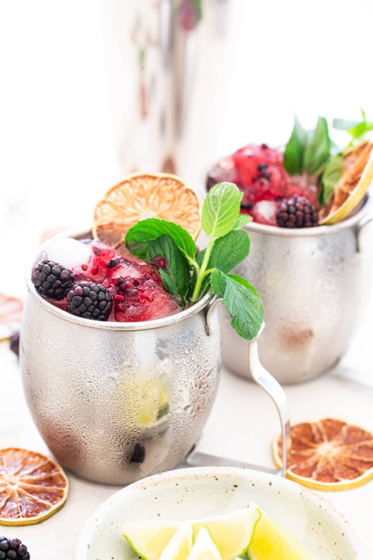 Two metal mugs filled with crushed ice, blackberries, mint, and dried lime slices—perfect for a refreshing Blackberry Moscow mule or any recipe with ginger beer. Lime wedges rest invitingly nearby for garnish.