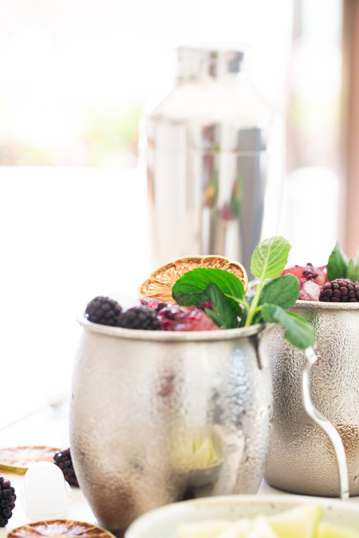 A silver cup with blackberries, mint, and a dried orange slice hints at a delicious blackberry cocktail recipe; a cocktail shaker is in the background.