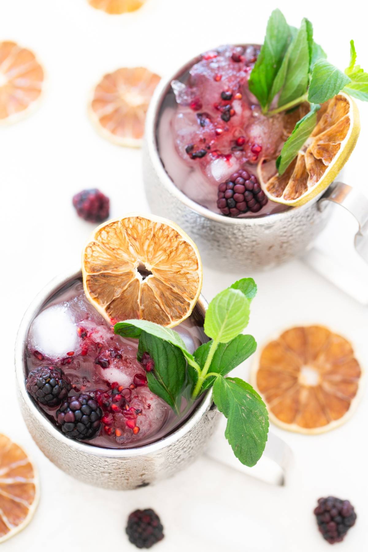 Two silver mugs with icy Blackberry Moscow mule cocktails, garnished with mint, blackberries, and dried orange slices—an irresistible recipe with ginger beer.