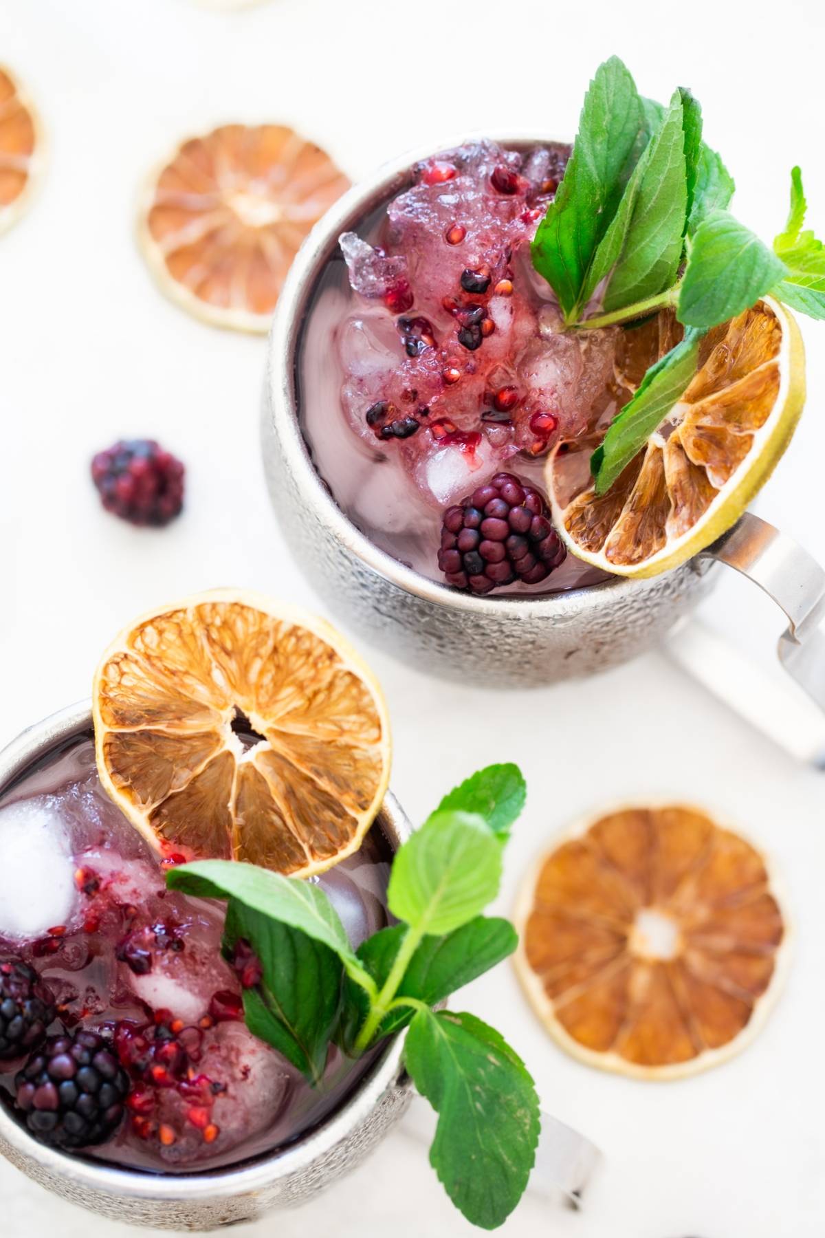 Two metal mugs with ice, blackberries, mint leaves, and dried citrus slices make the perfect Blackberry Moscow mule, surrounded by dried citrus for a refreshing cocktail with vodka.