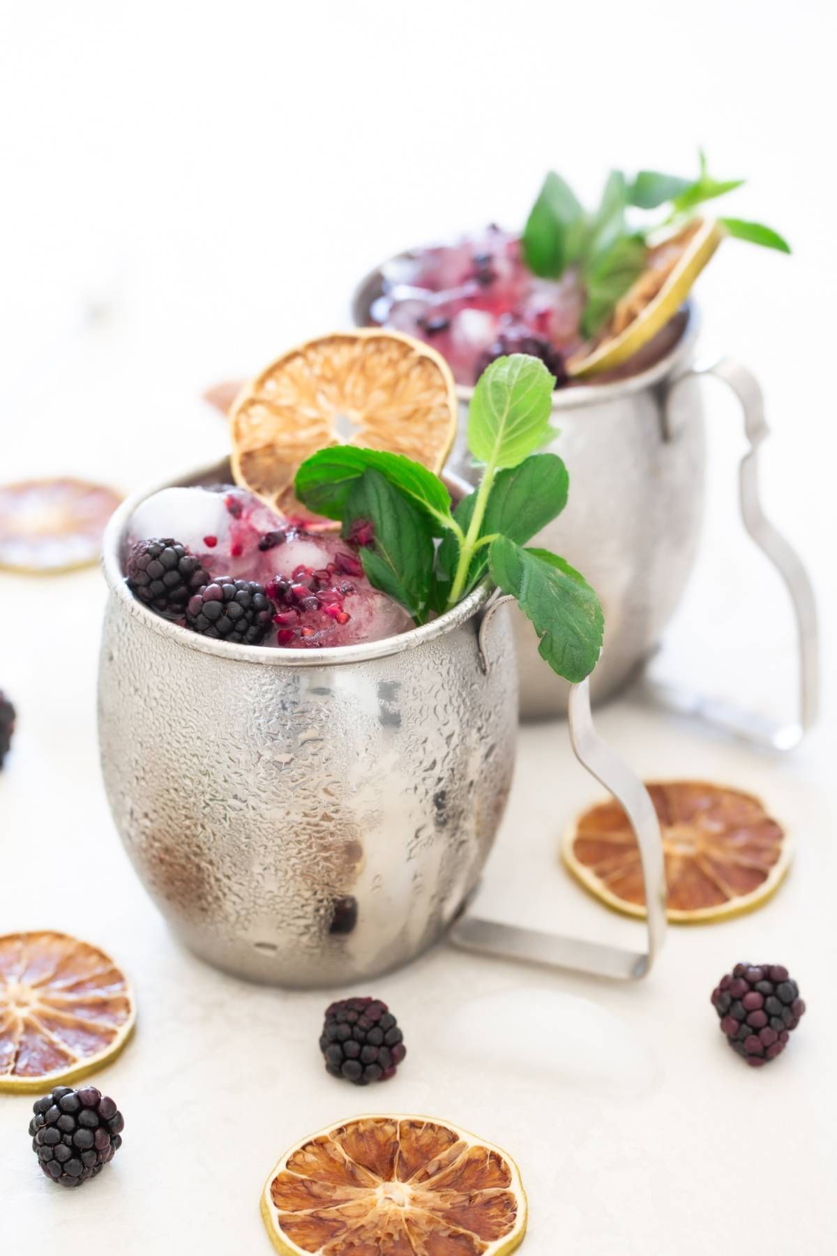 Two metal mugs filled with ice, blackberries, mint, and dried citrus slices hint at a refreshing Blackberry Moscow mule—perfect if you’re craving a recipe with ginger beer. Dried orange rounds complete this vibrant scene.