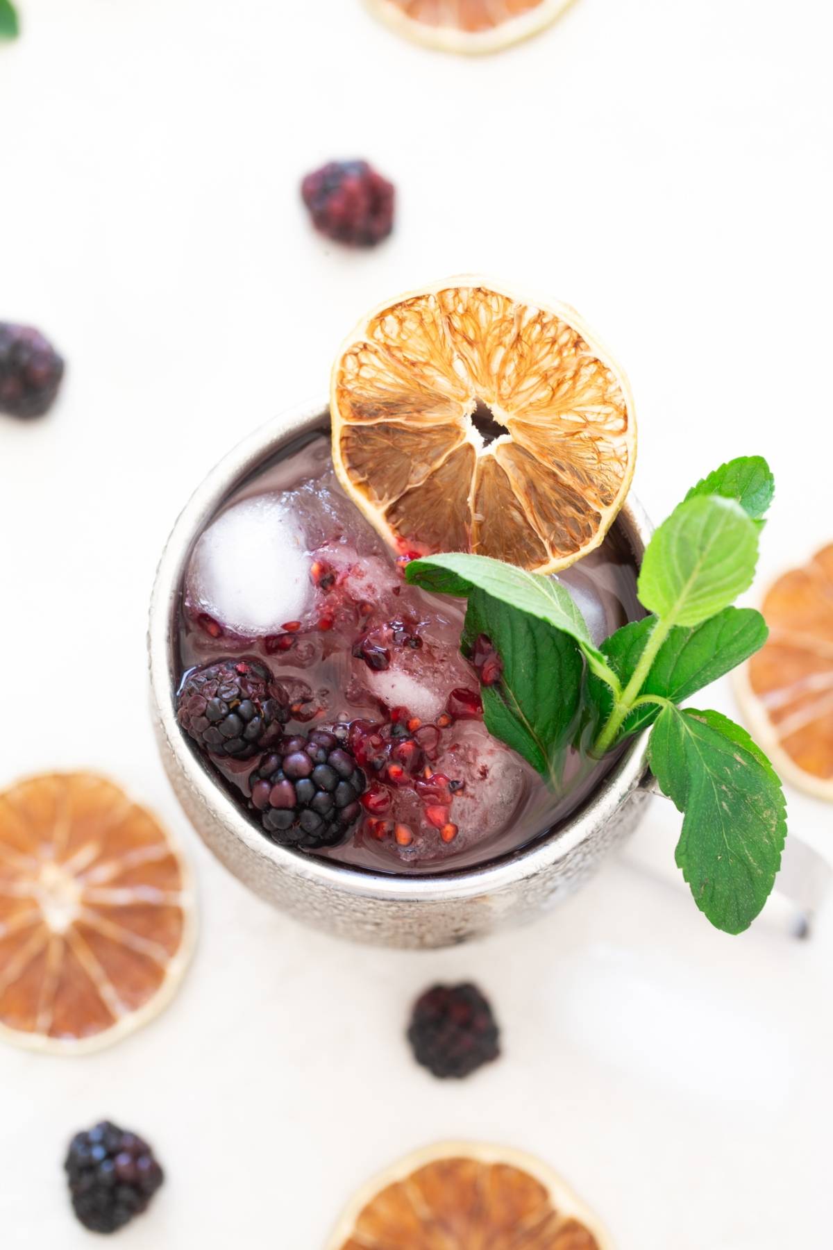 A refreshing blackberry cocktail recipe served in a silver mug with ice, mint, and a dried orange slice garnish—perfect for those looking for a unique recipe with ginger beer. Surrounded by fresh fruit, it's a vibrant twist on classic cocktails.
