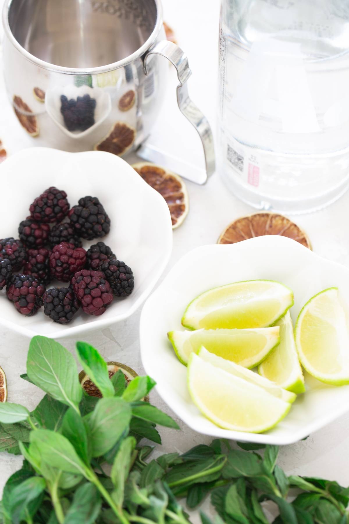 Fresh blackberries, lime wedges, mint leaves, a metal mug, and a bottle of clear liquid on a white surface—perfect ingredients for a cocktail with vodka or trying out a recipe with ginger beer.