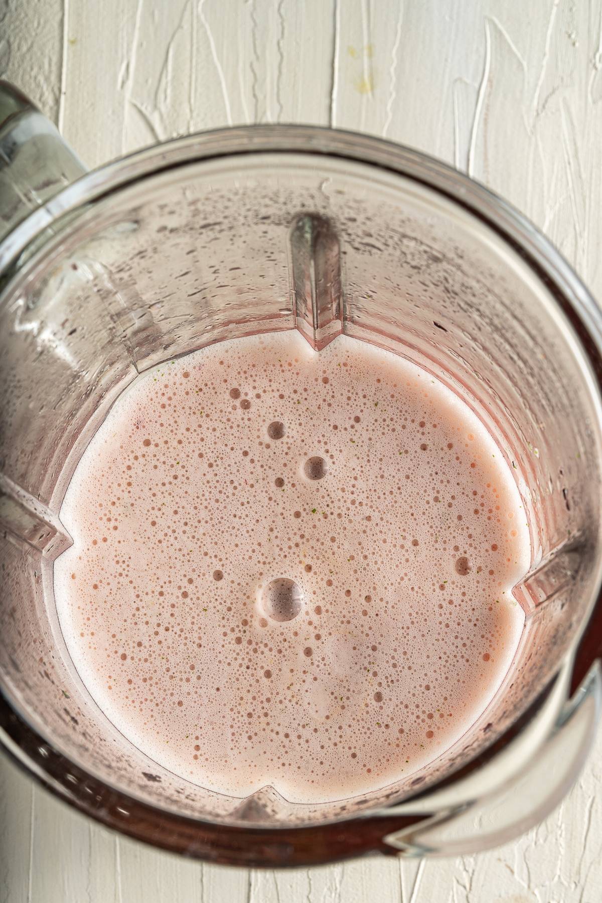 Pink smoothie with bubbles inside a clear blender jar on a white textured surface—a refreshing summer treat inspired by citrus and blueberry flavours, perfect for pairing with your favourite blueberry popsicle recipe.