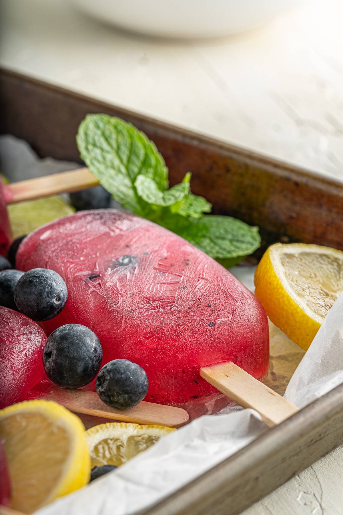 Red popsicle with blueberries, lemon slices, and mint leaves on a tray with parchment paper—this refreshing summer treat is inspired by a Blueberry Lemonade Popsicles recipe.