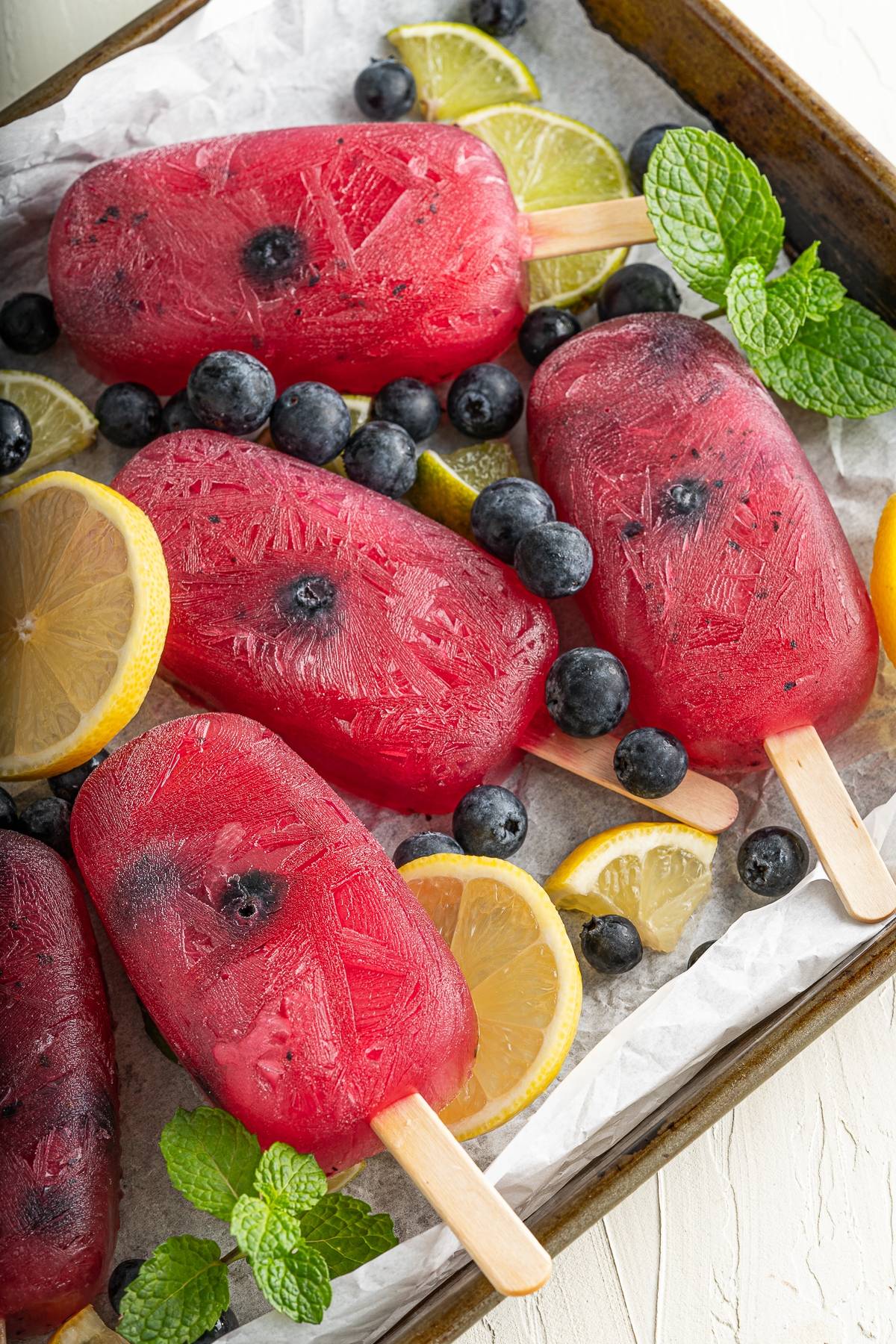 Red popsicles with blueberries, lemon slices, lime wedges, and mint leaves on a tray with parchment paper—a refreshing summer treat perfect for those seeking a blueberry popsicle recipe without yogurt.