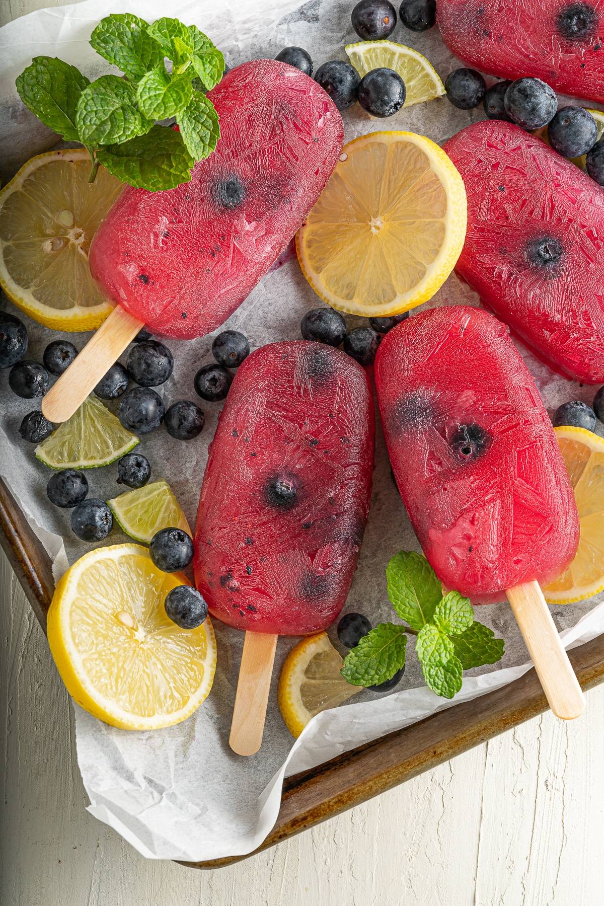 Blueberry Lemonade Popsicles with blueberries, lemon slices, lime wedges, and mint leaves rest on a tray lined with parchment paper—a refreshing summer treat perfect for any hot day.
