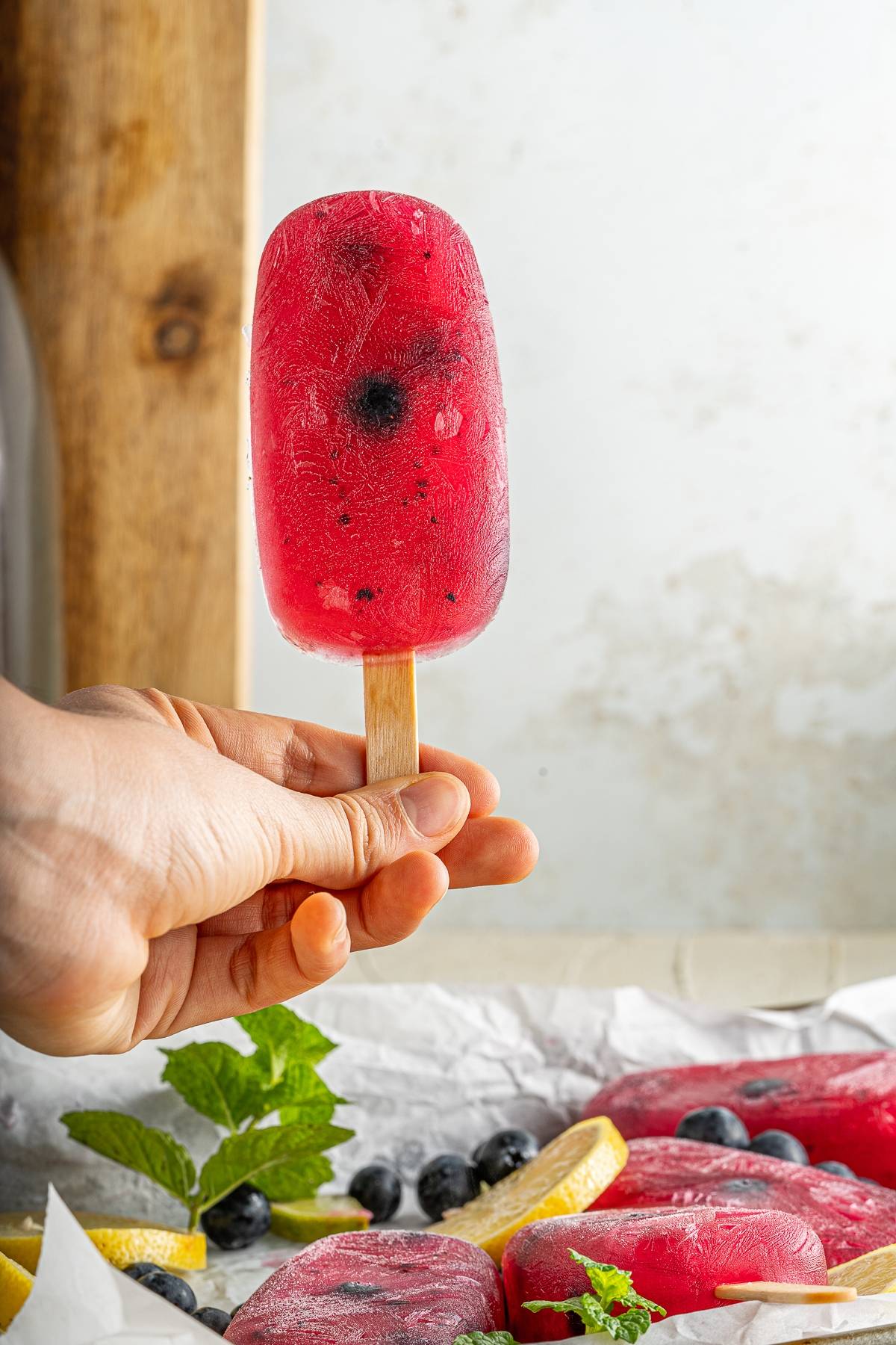 Hand holding a citrus and blueberry flavoured popsicle above a tray with more Blueberry Lemonade Popsicles, blueberries, lemon slices, and mint.