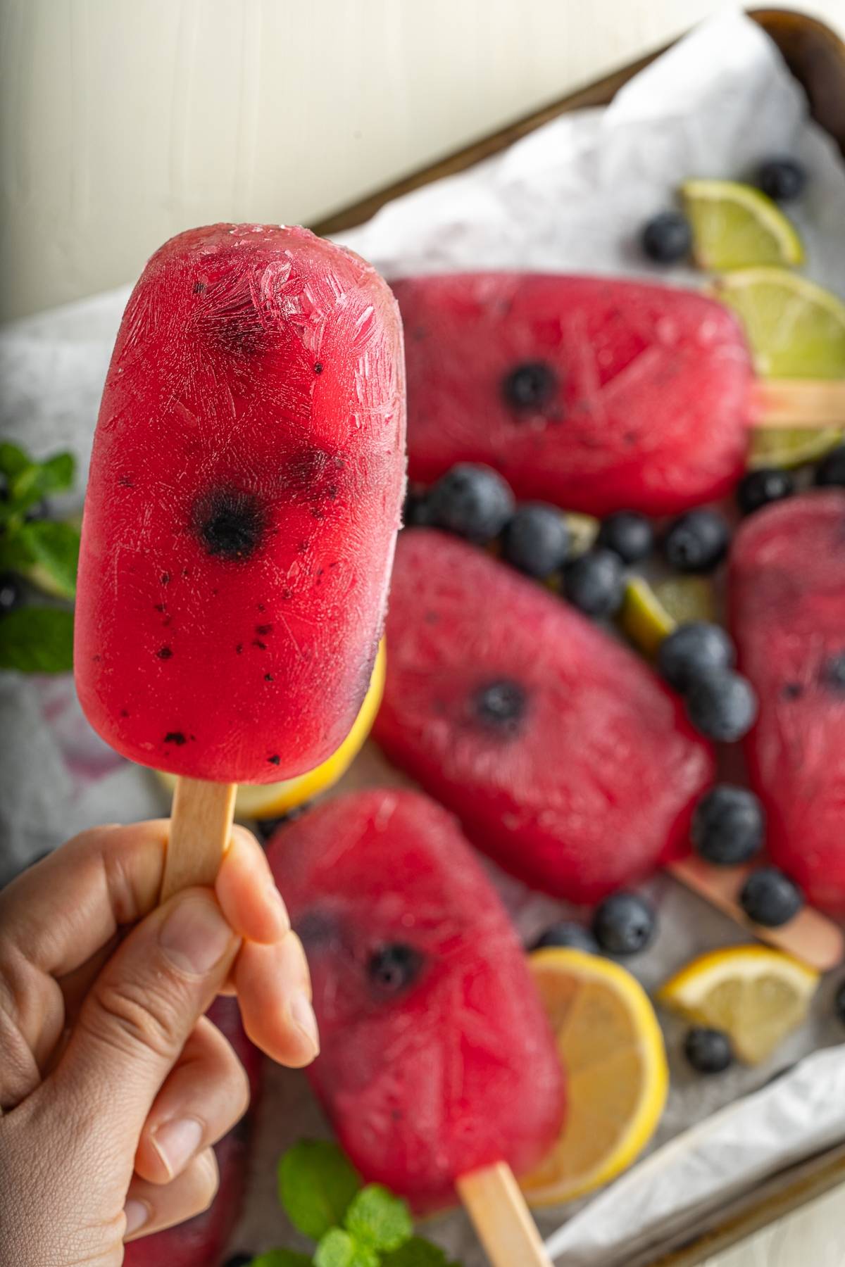 Hand holding a red popsicle with blueberries; more Blueberry Lemonade Popsicles, lemon slices, and blueberries in the background—a refreshing summer treat perfect for anyone seeking a delicious blueberry popsicle recipe.