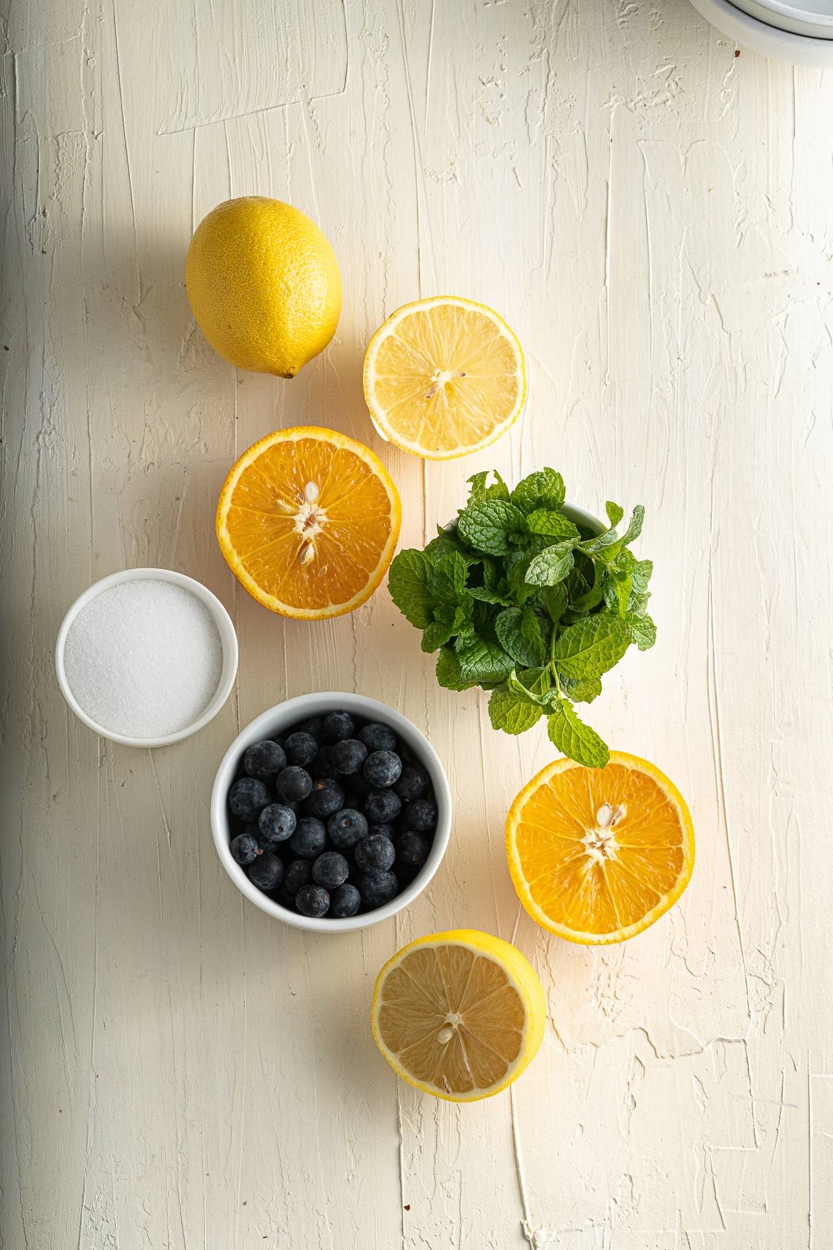 Lemon, orange halves, blueberries, fresh mint, and a bowl of sugar on a white textured surface—perfect ingredients for a refreshing summer treat or your next citrus and blueberry flavoured popsicle.