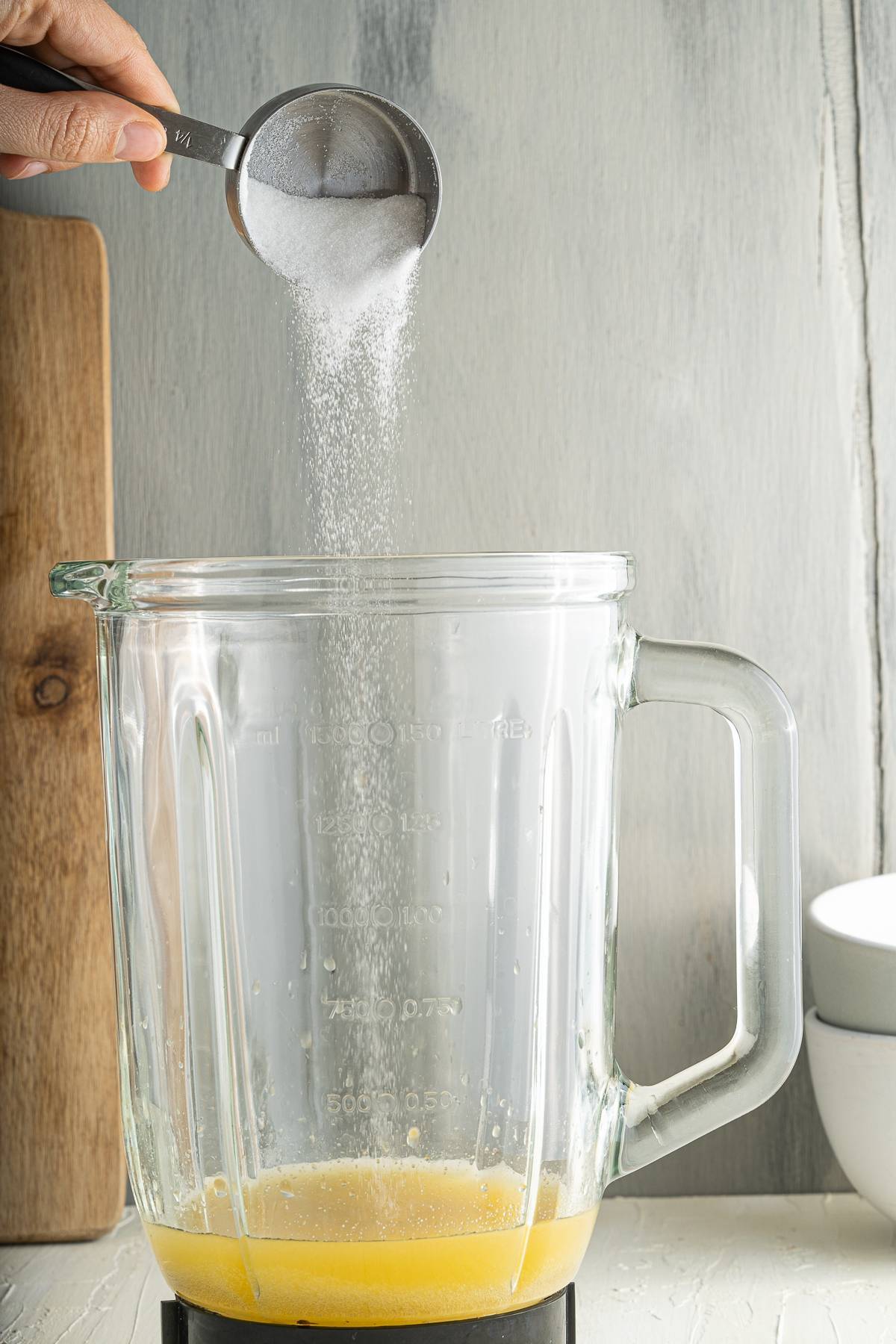 A hand pours sugar from a metal measuring cup into a glass blender with liquid at the bottom, preparing the base for citrus and blueberry flavoured popsicles.