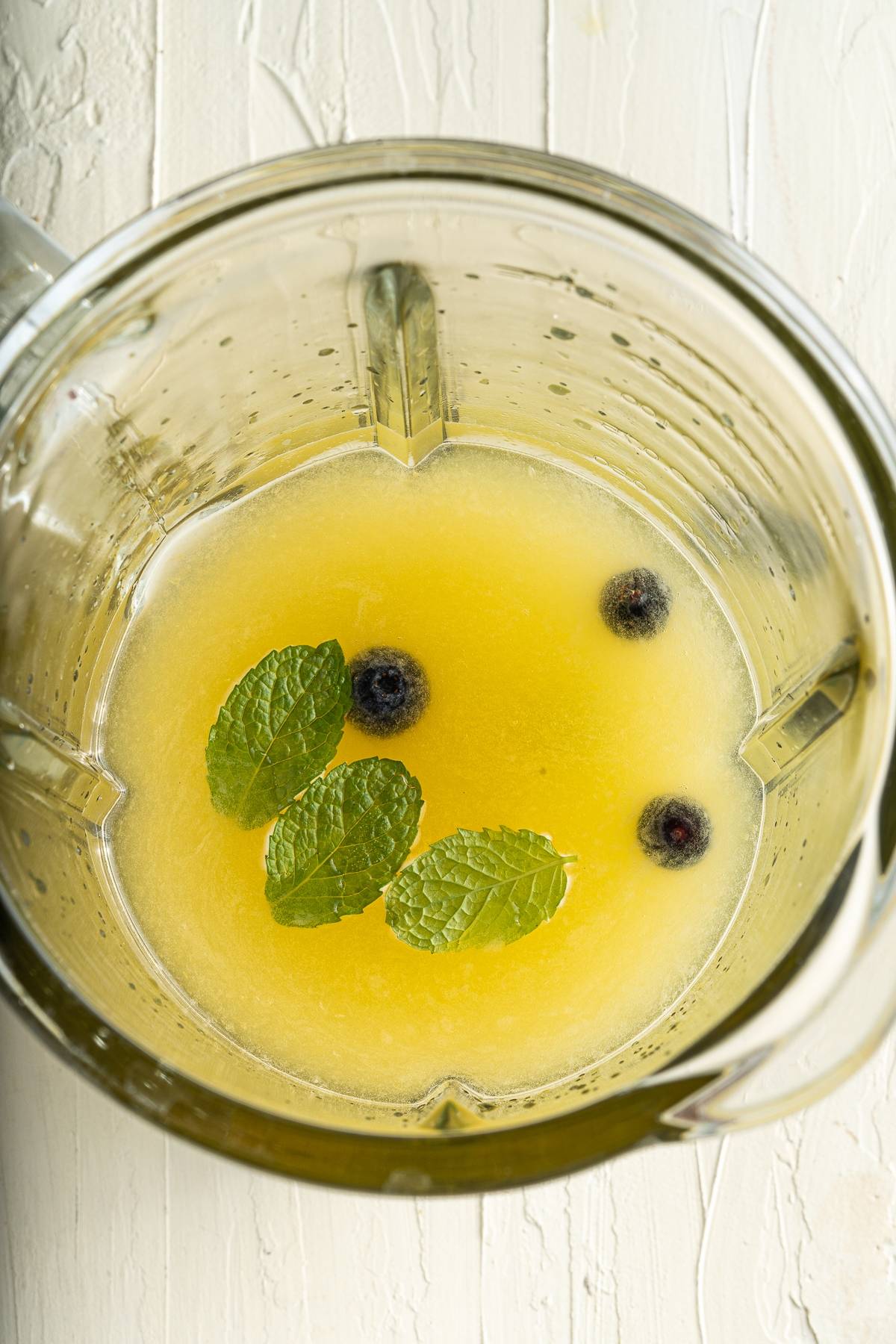 A blender with yellow juice, mint leaves, and blueberries on a white wooden surface—perfect ingredients for Blueberry Lemonade Popsicles, a refreshing summer treat.