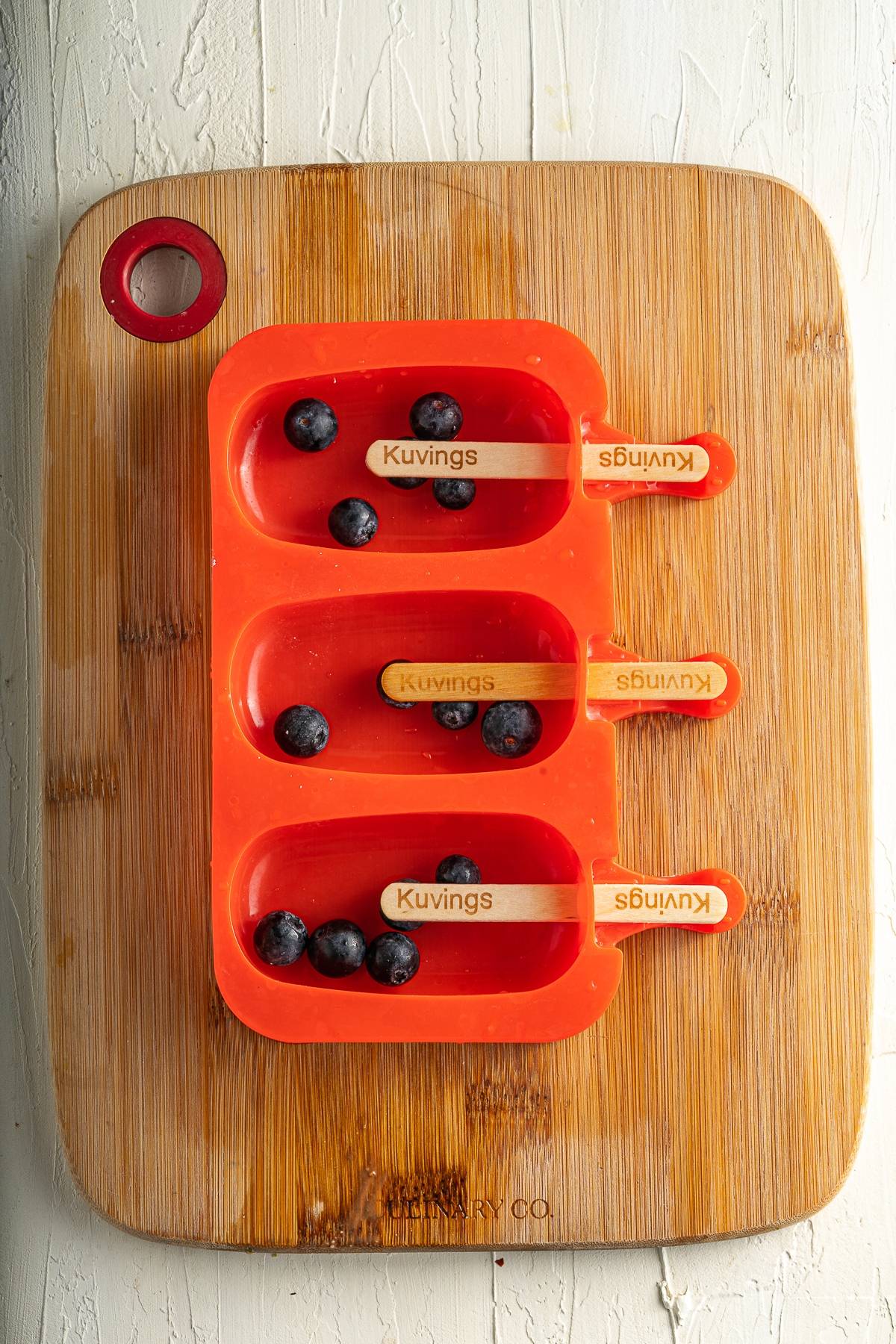 Three orange popsicle molds with wooden sticks and blueberries inside, on a wooden cutting board—perfect for making blueberry lemonade popsicles or trying your favorite blueberry popsicle recipe without yogurt.