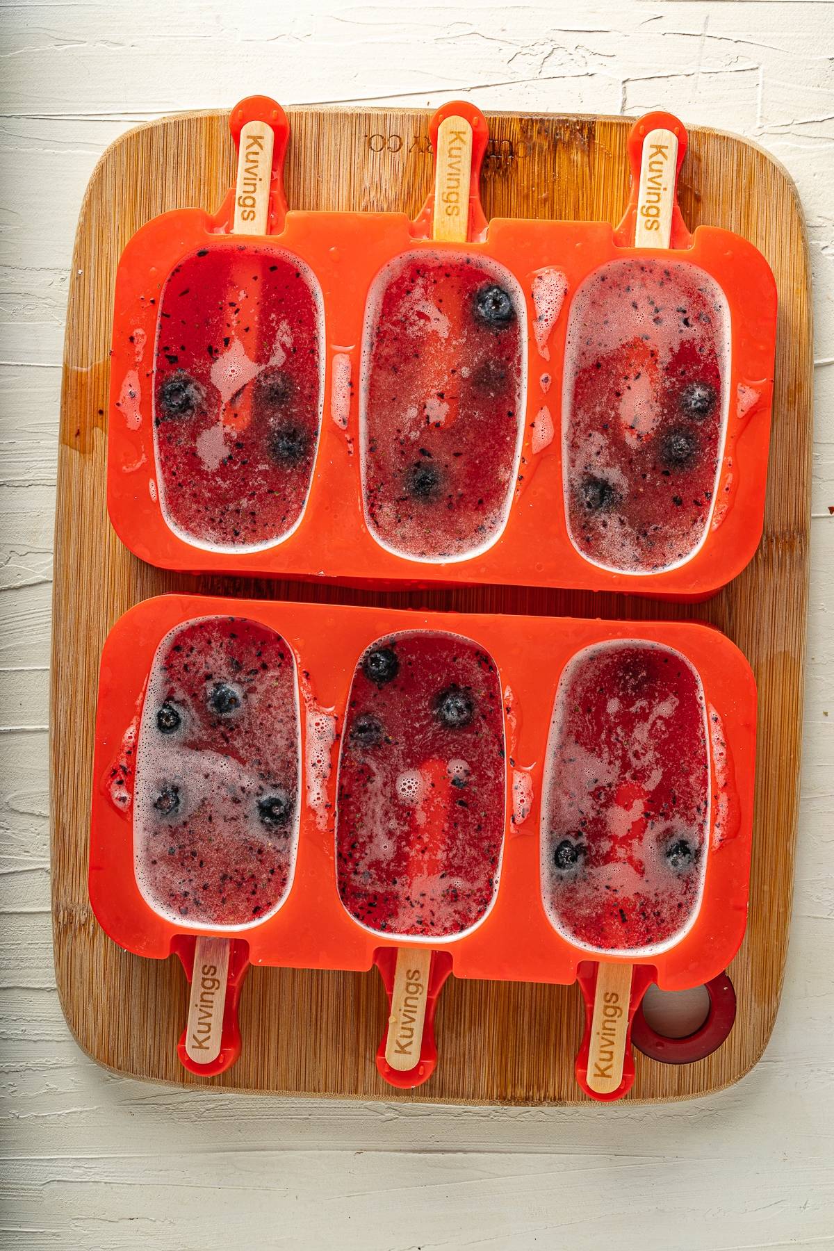 Six homemade citrus and blueberry flavoured popsicles in orange silicone molds on a wooden board, with visible blueberries inside.