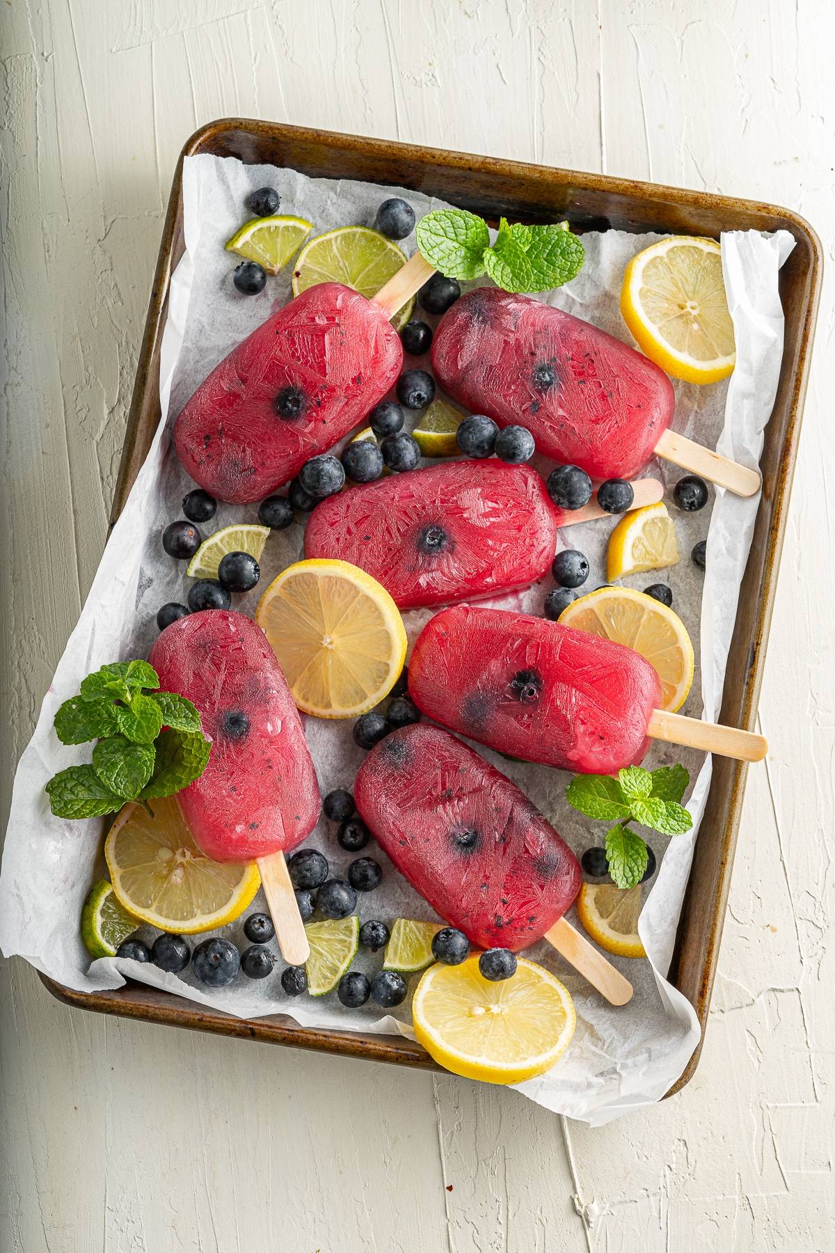 Six refreshing summer treat popsicles on a tray, with citrus and blueberry flavoured popsicles surrounded by blueberries, lemon and lime slices, and fresh mint leaves.