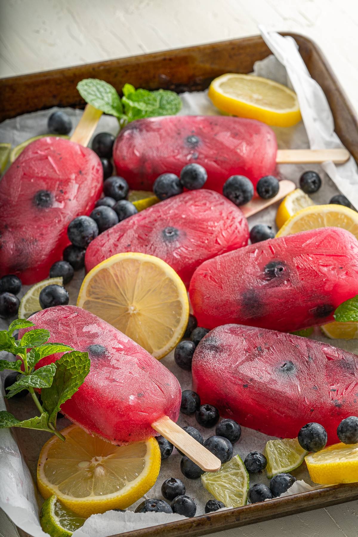 Six refreshing summer treat berry popsicles with citrus and blueberry flavours, blueberries, lemon slices, and mint leaves arranged on a tray with parchment paper.