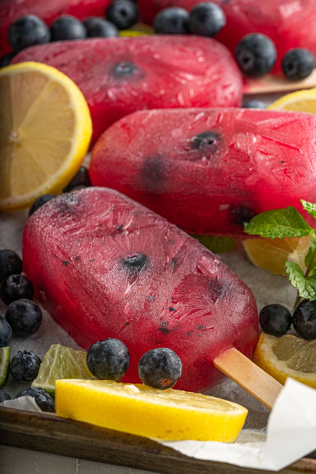 Bright red Berry Blueberry Lemonade Popsicles with blueberries, lemon slices, and mint leaves on a tray—an irresistibly refreshing summer treat.