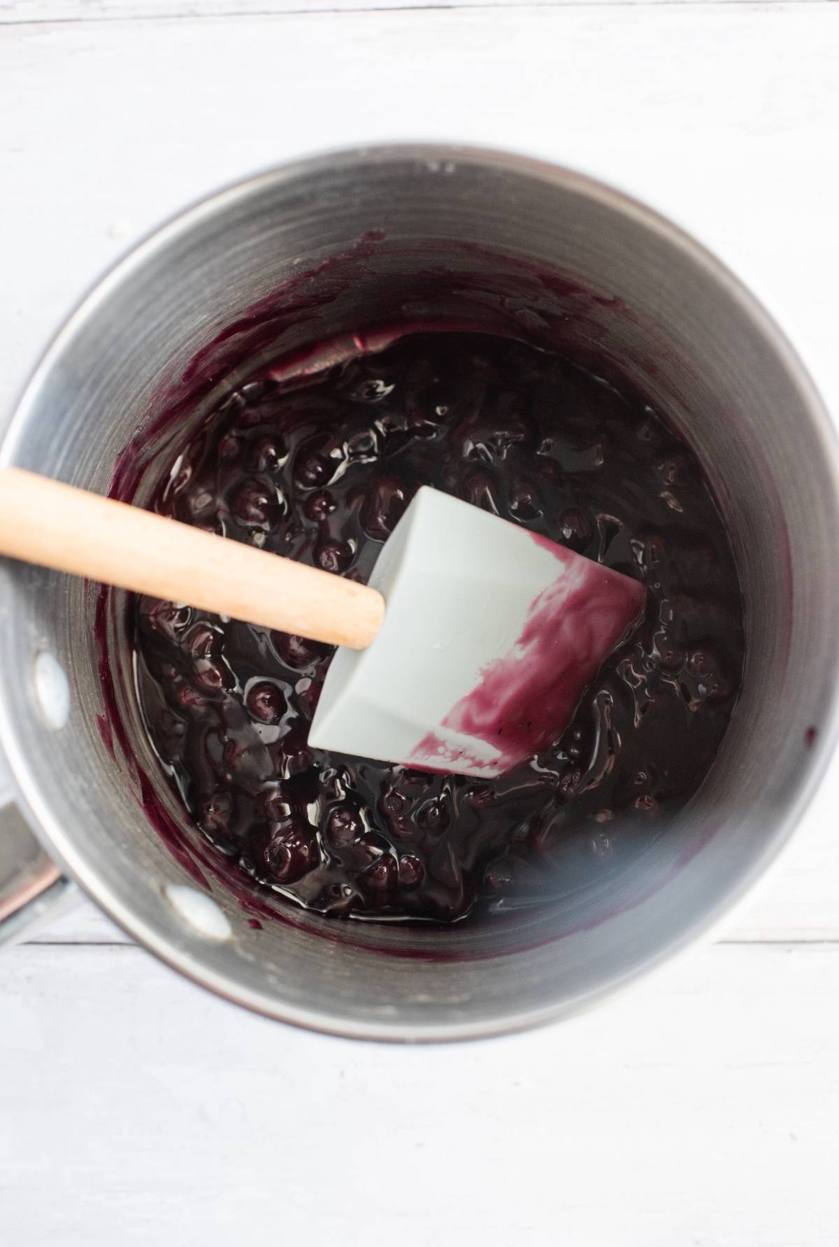 A saucepan with dark berry sauce, perfect for a Blueberry Pie Meringue Squares filling, and a spatula mixing the contents on a light surface.